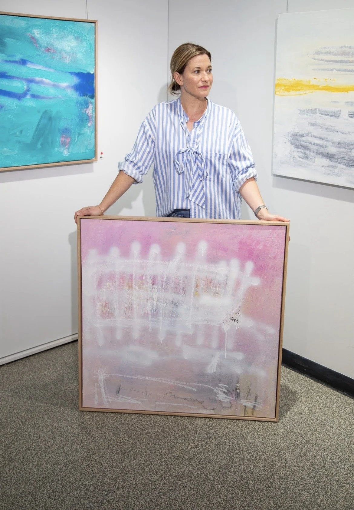 A woman in a striped shirt standing in an art gallery, holding a pink and white abstract painting with light background, with additional abstract paintings on the white walls behind her.