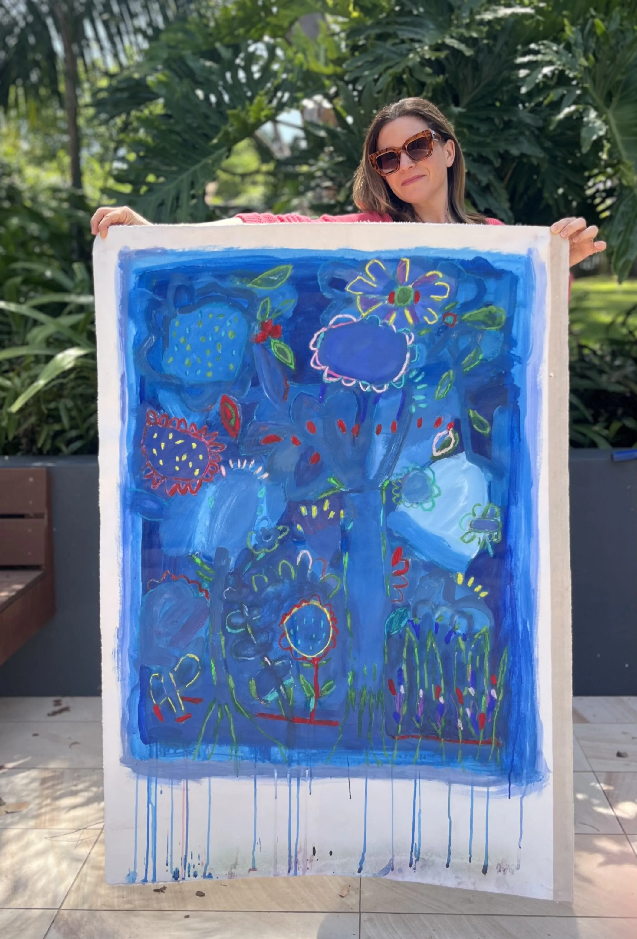 A woman with brown hair, wearing sunglasses and a red shirt, holding a colorful abstract painting, outdoors with green foliage in the background.