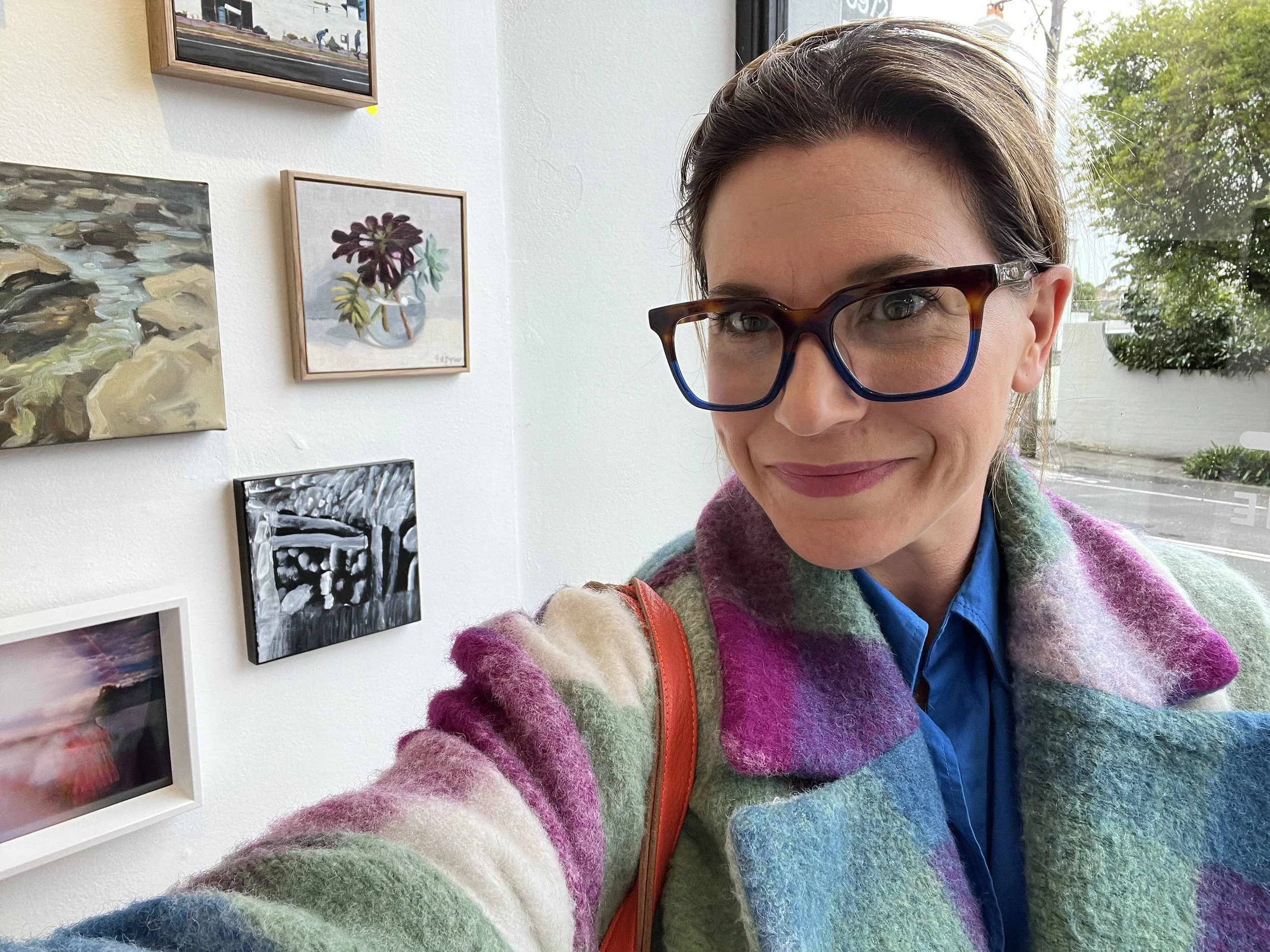 A woman wearing large tortoiseshell glasses and a multicolored fuzzy sweater, taking a selfie in an art gallery with paintings on the wall.