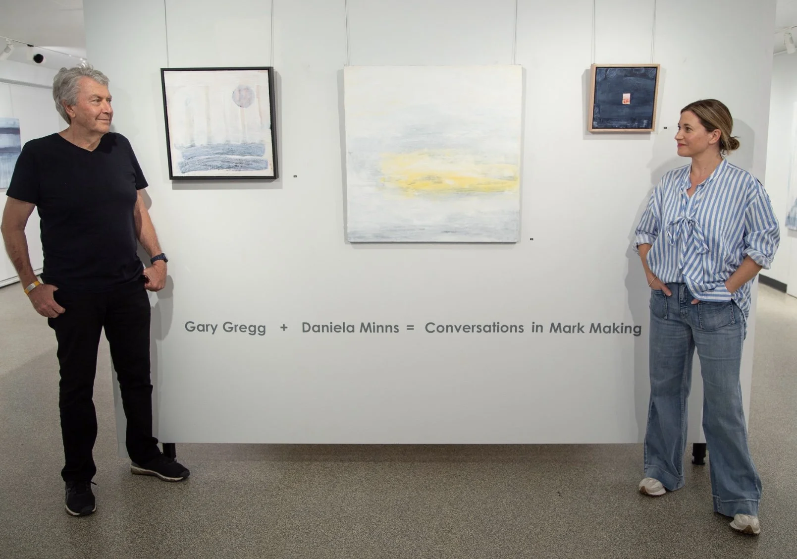 Two people standing in an art gallery, looking at paintings. The man on the left has gray hair, is wearing a black t-shirt and dark pants. The woman on the right has brown hair, is wearing a blue and white striped shirt and jeans. There are four pain