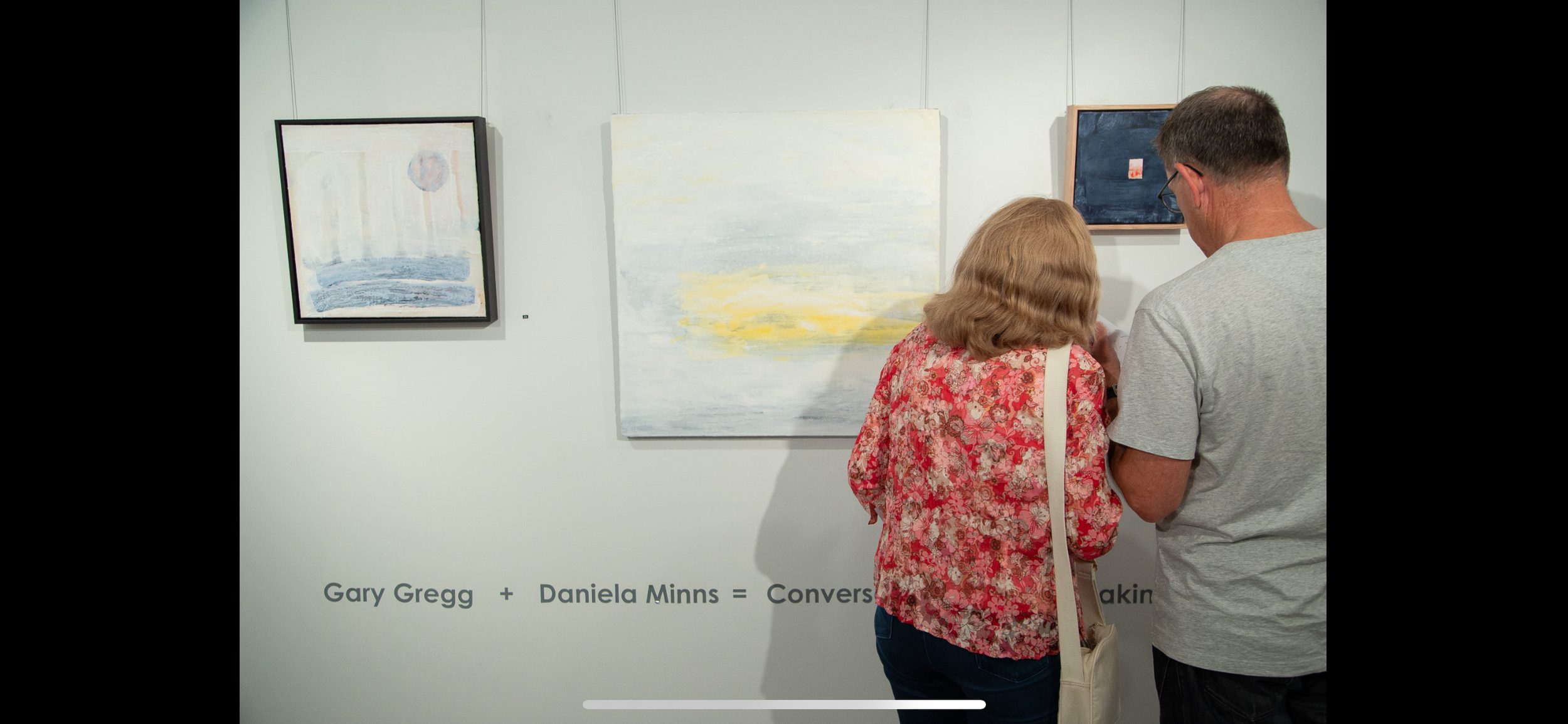 Two people looking at abstract paintings in an art gallery. The wall has a text that reads 'Gary Gregg + Daniela Minns = Conversation'.