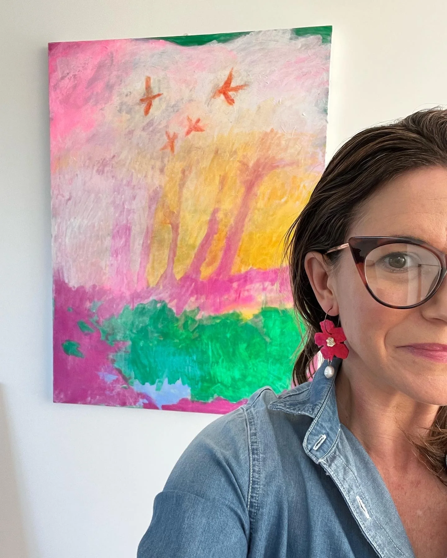 A woman wearing glasses and a blue denim shirt with a pink flower earrings, taking a selfie in front of a colorful abstract painting with shades of pink, yellow, green, and purple, with birds in flight depicted in the artwork.