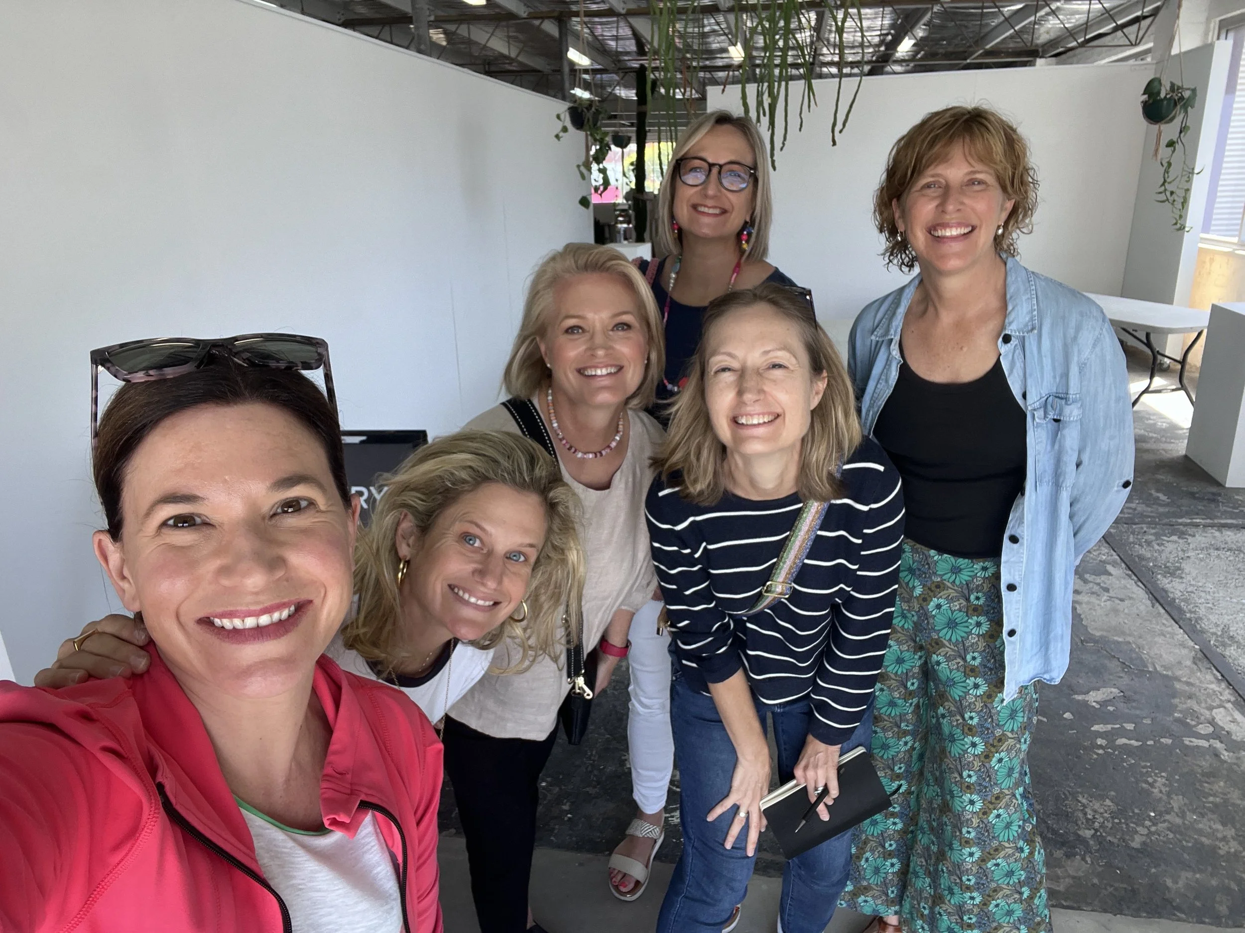 A group of smiling women taking a selfie in an indoor space with high ceilings and hanging plants.