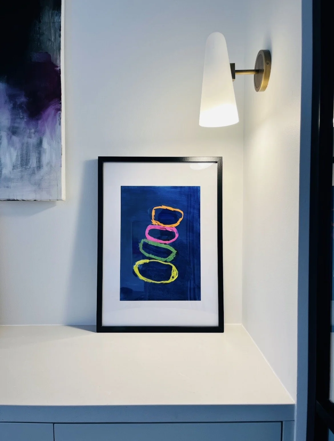 Framed abstract artwork with colorful ovals on a dark blue background, placed on a white surface, with a wall-mounted white lamp above illuminating the artwork.