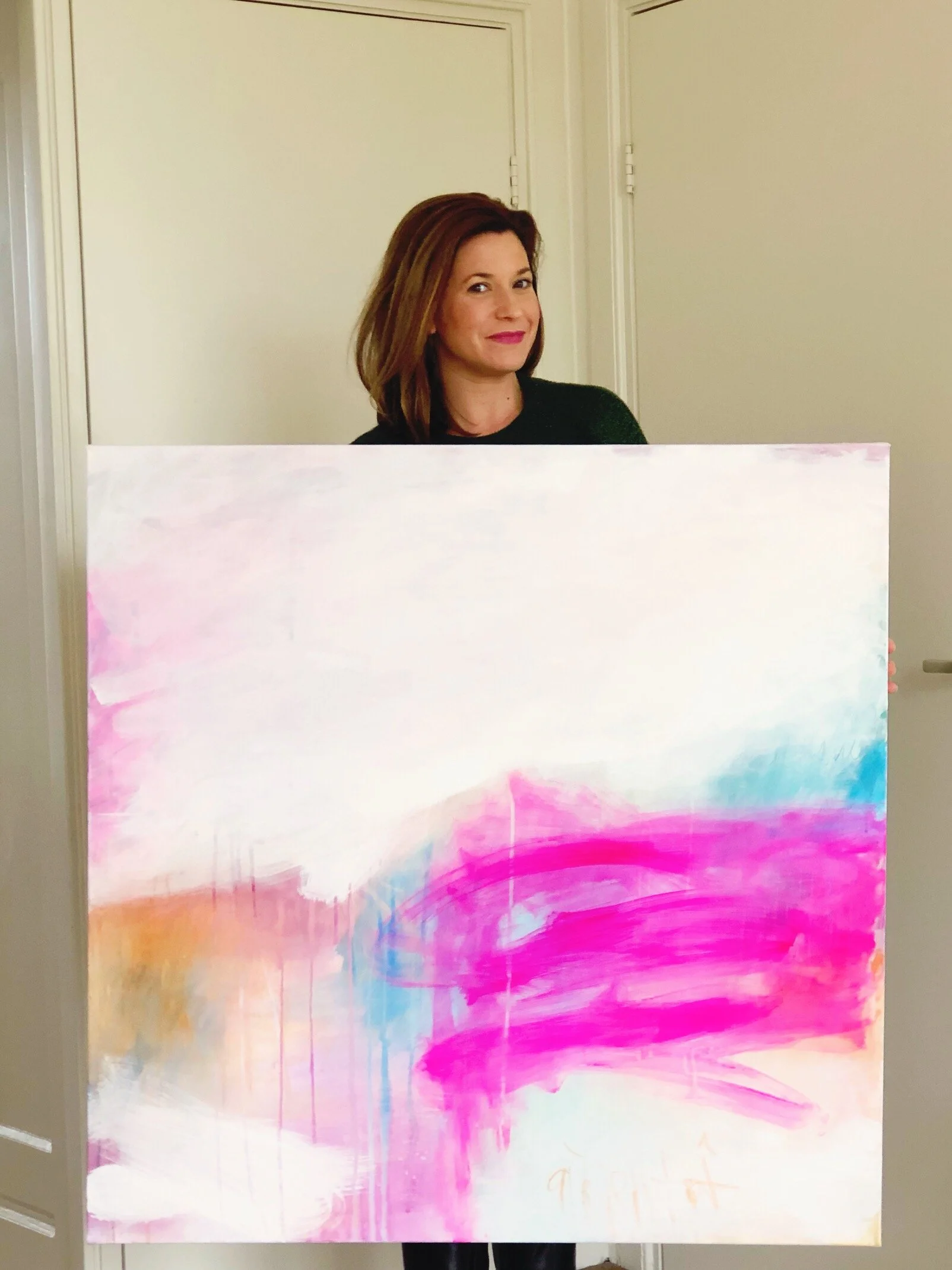 A woman with shoulder-length brown hair and a black shirt holding an abstract painting with pink, purple, blue, and orange colors.