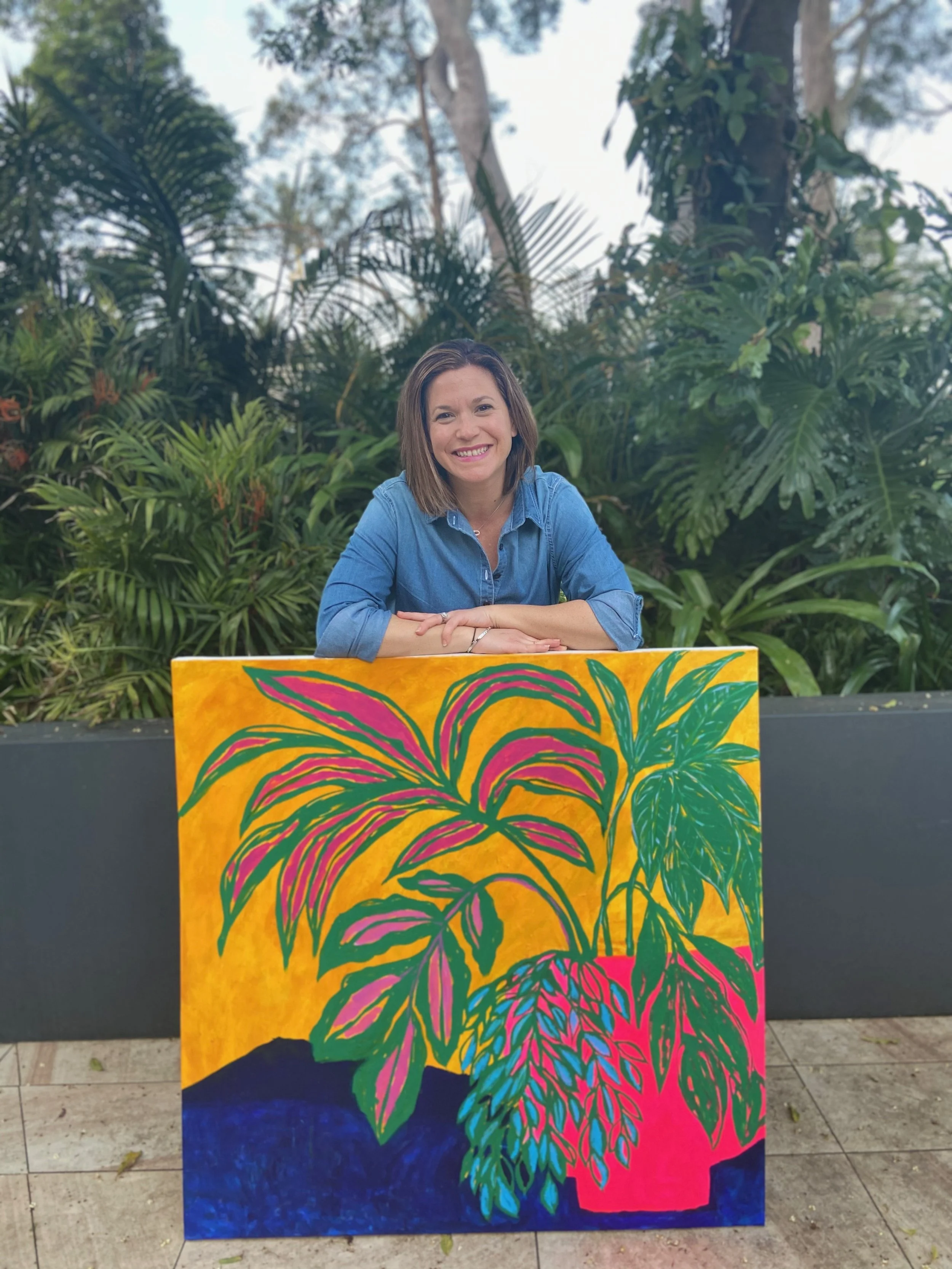 A woman with shoulder-length brown hair, wearing a blue denim shirt, is sitting at a table behind a colorful abstract painting of a large plant.