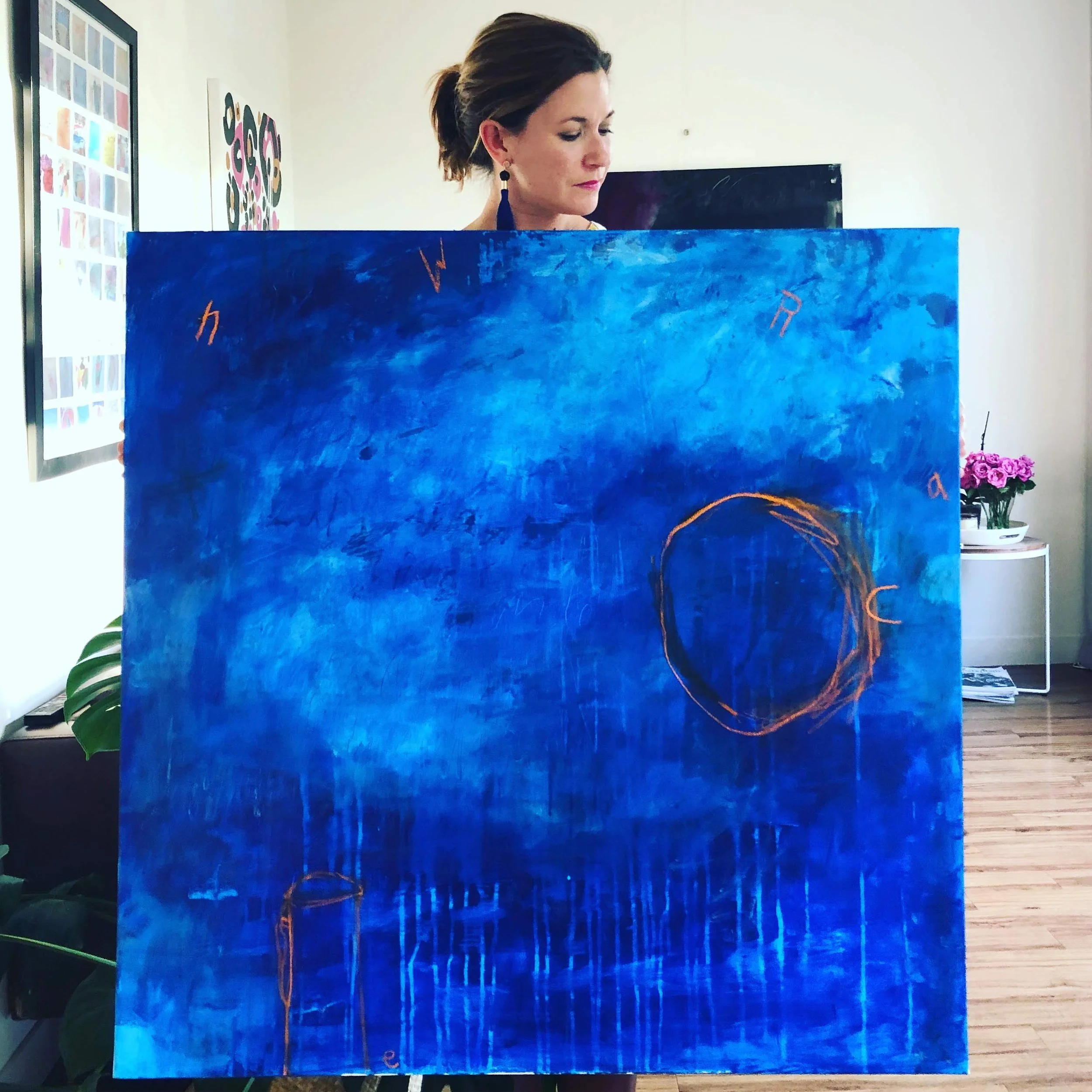 A woman with brown hair and earrings looking down behind a large, abstract blue-painted canvas with orange squiggles.