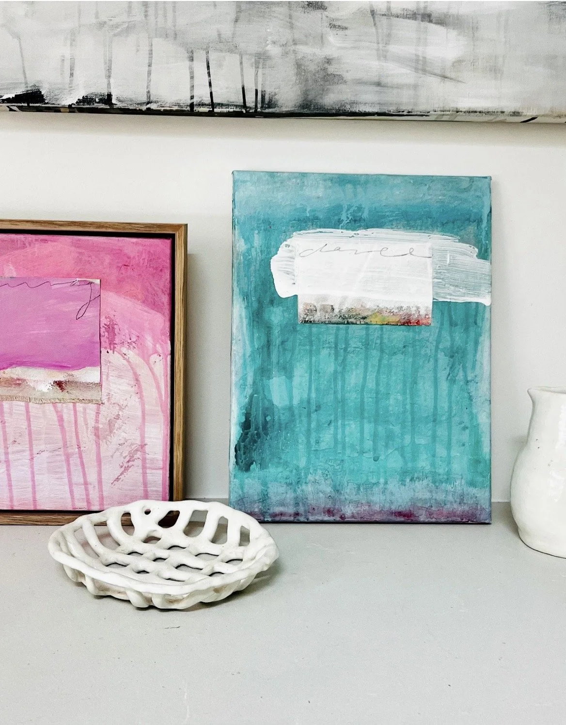 Two abstract paintings leaning against a white wall, one pink with a wooden frame and the other teal with a white painted rectangle, a white ceramic bowl with an open lattice design, and a white ceramic vase on a surface.