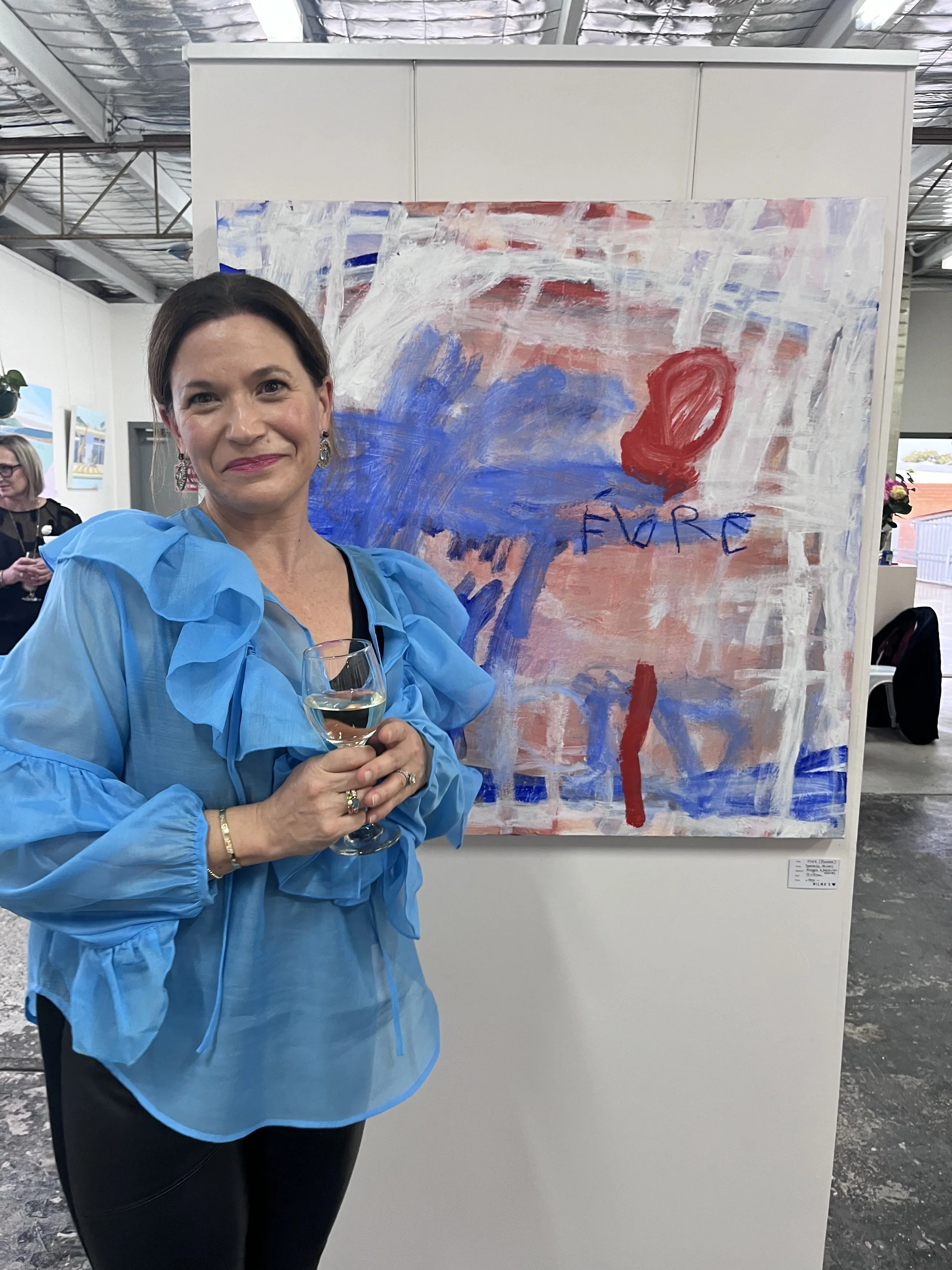 A woman in a blue blouse holding a glass of white wine at an art exhibition. Behind her is an abstract painting with the word 'FORCE' written on it.