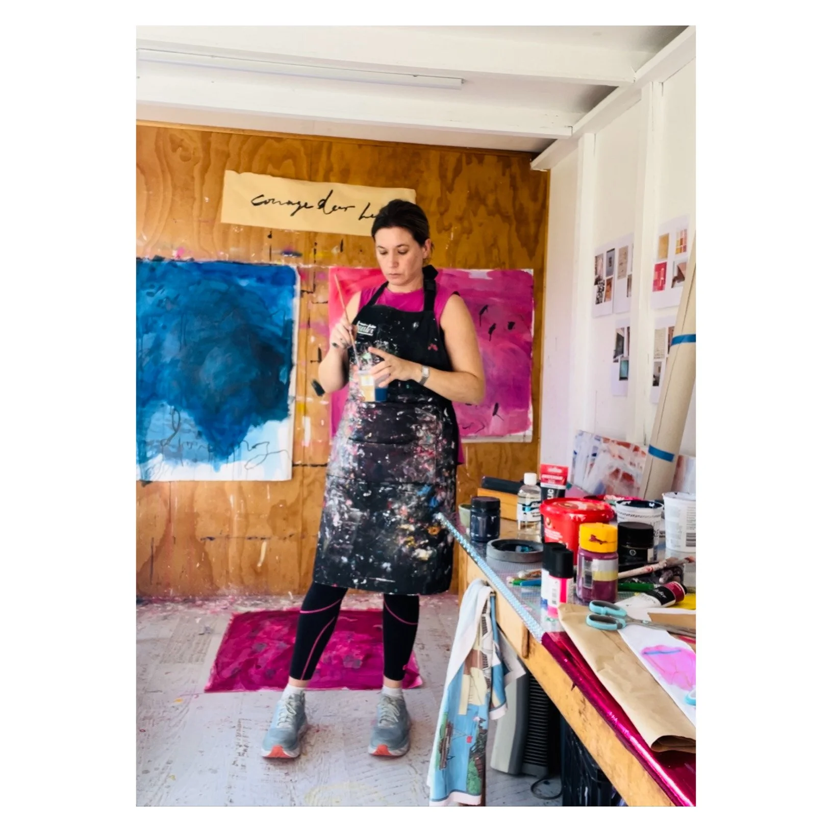 A woman with short dark hair wearing paint-splattered apron and black leggings with pink lines inside an art studio, standing on a red rug in front of abstract paintings on a wooden wall, with a table filled with art supplies including paint jars, sc