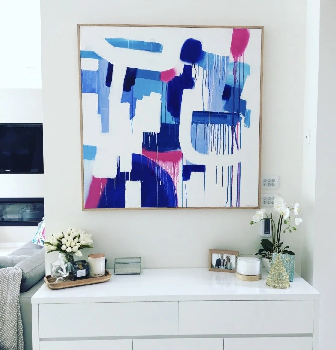 Decorative white sideboard with a floral arrangement, candles, and framed photos, with an abstract blue, pink, and white painting hanging above.