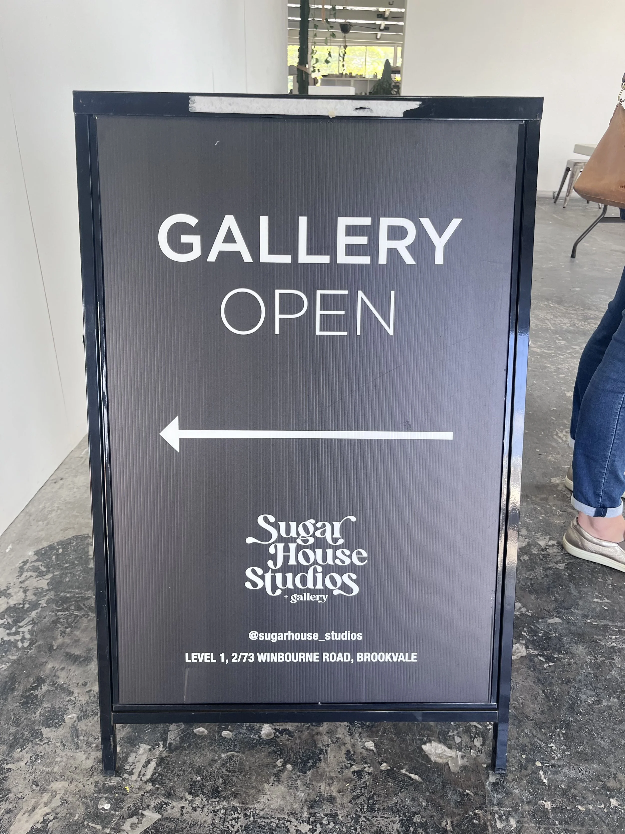 Freestanding black sign with white text indicating that the Sugarhouse Studios gallery is open, with an arrow pointing to the left, and including location address at Winbourne Road, Brookvale.