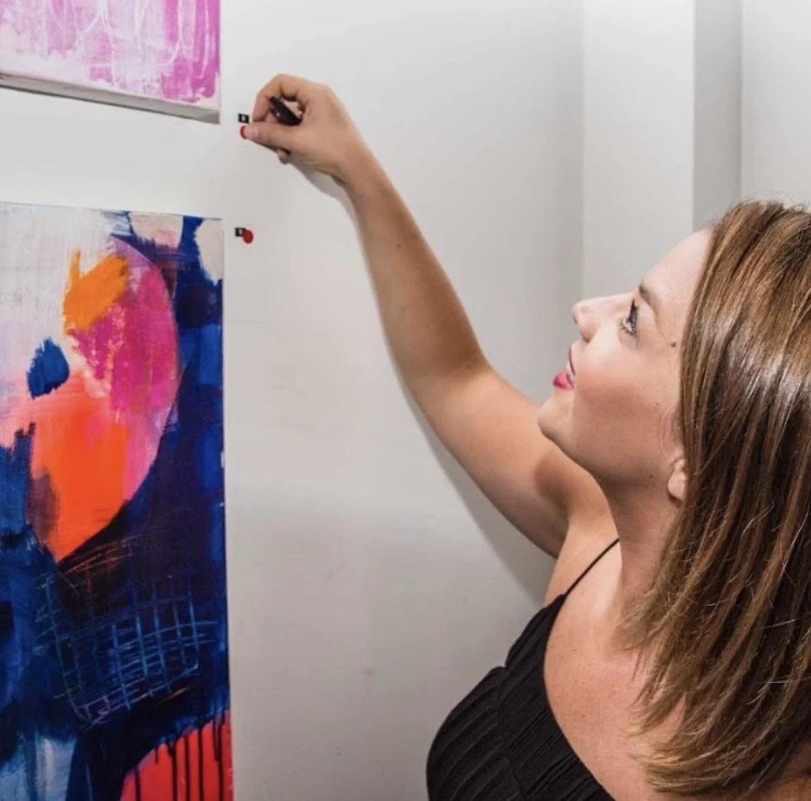 A woman with brown hair and a black top hanging a colorful abstract painting on a white wall.
