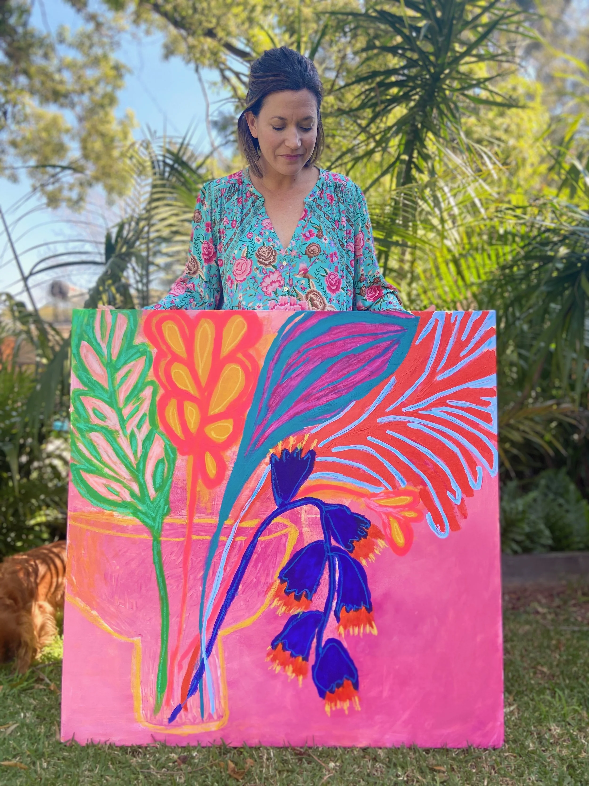 A woman in a floral patterned blouse standing outdoors behind a colorful painting with floral and abstract designs on a pink background.