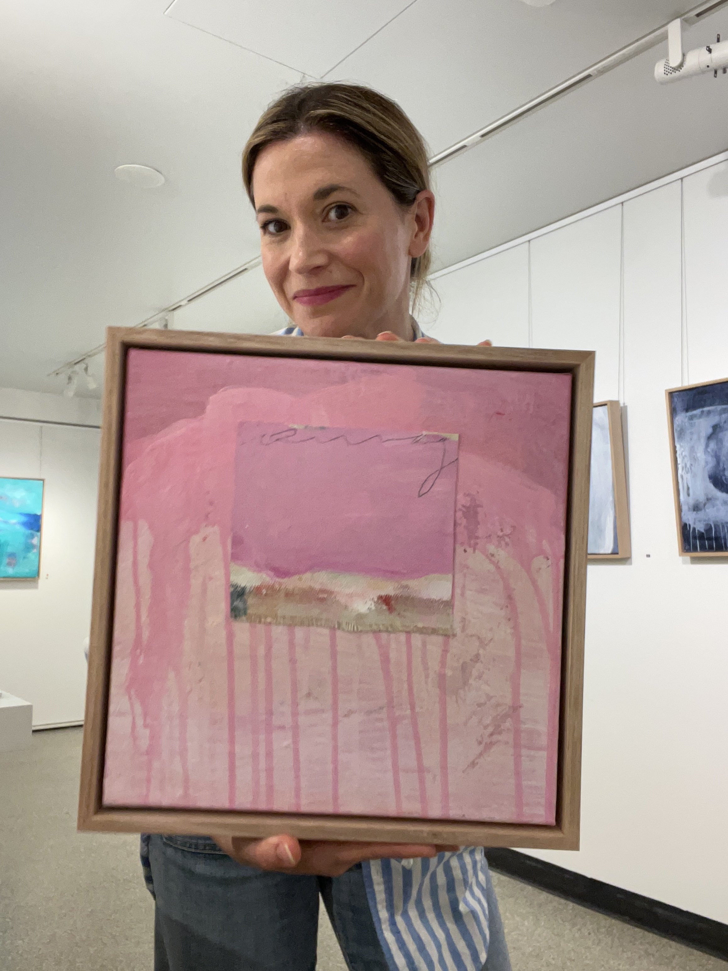 A person holding a framed pink abstract painting in an art gallery, with white walls and other framed artworks visible in the background.