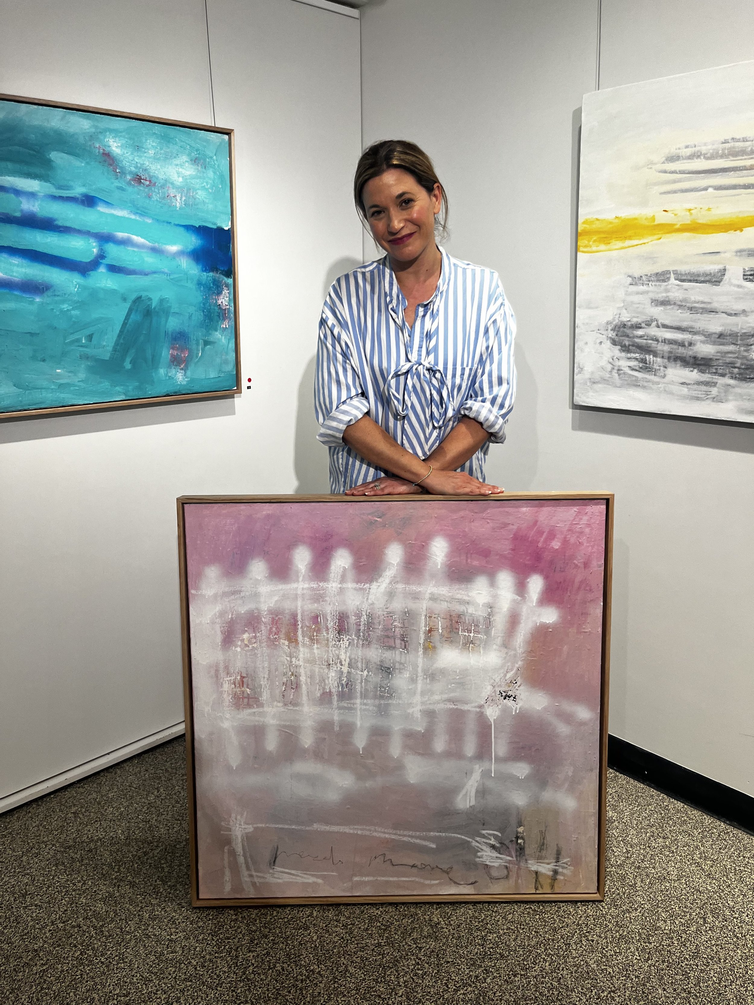 A woman in a blue and white striped shirt standing in an art gallery, surrounded by abstract paintings in shades of blue, pink, white, and yellow on the walls.
