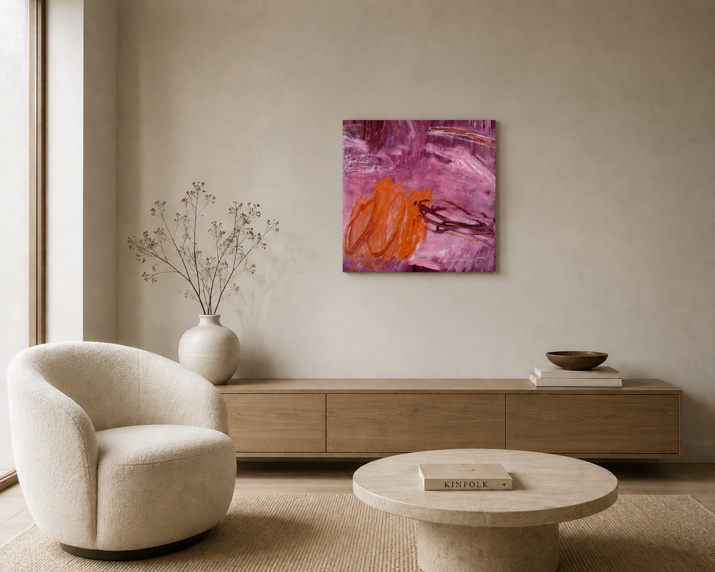 Modern living room with a beige armchair, a wooden sideboard, abstract pink and orange artwork on the wall, a white vase with dried branches, a round coffee table with a book titled 'KINFOLK', and a natural fiber rug.