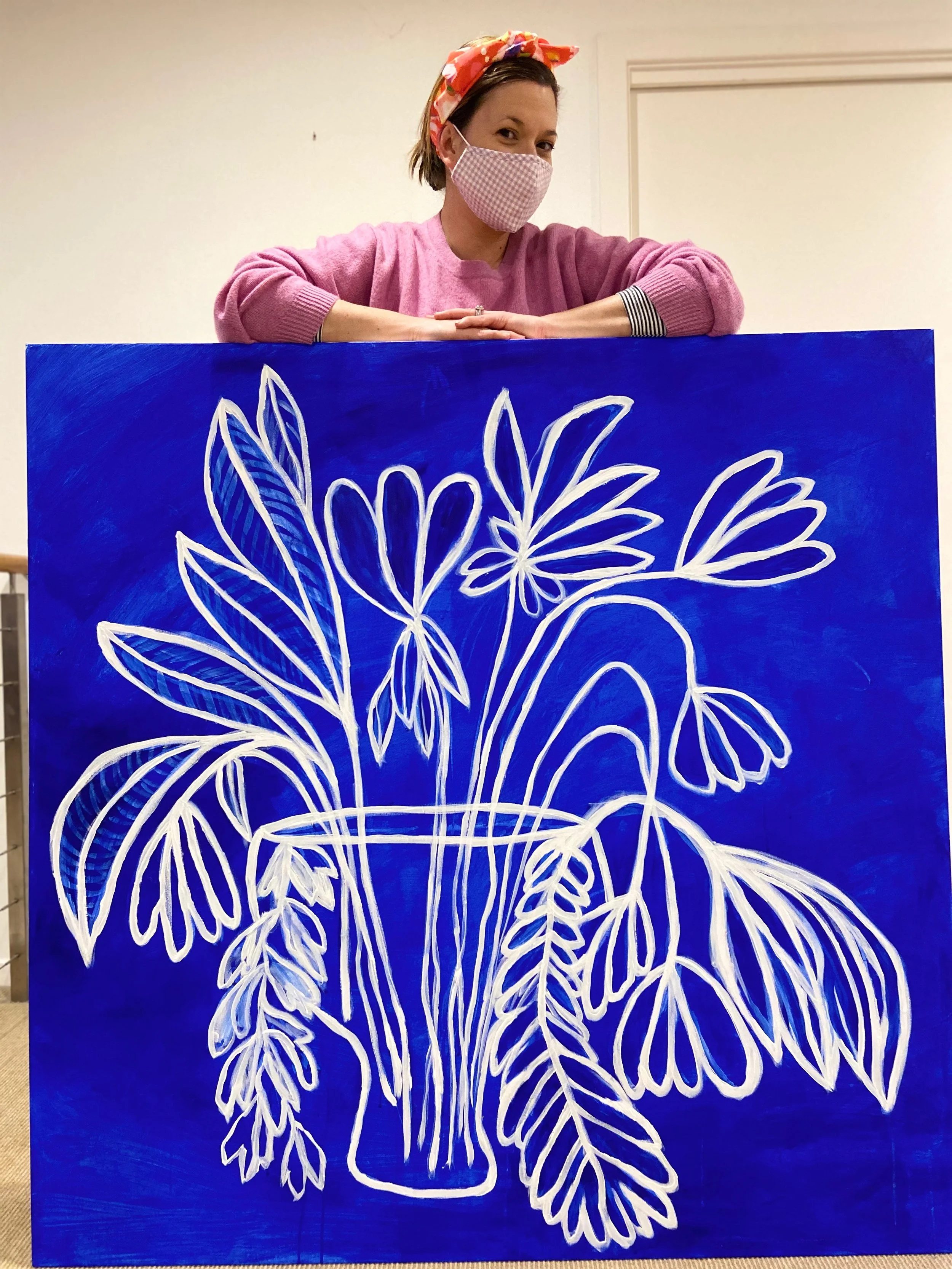 Woman wearing a pink sweater and a pink checkered face mask, with a red and pink patterned headscarf, standing behind a blue painting of white flower outlines.