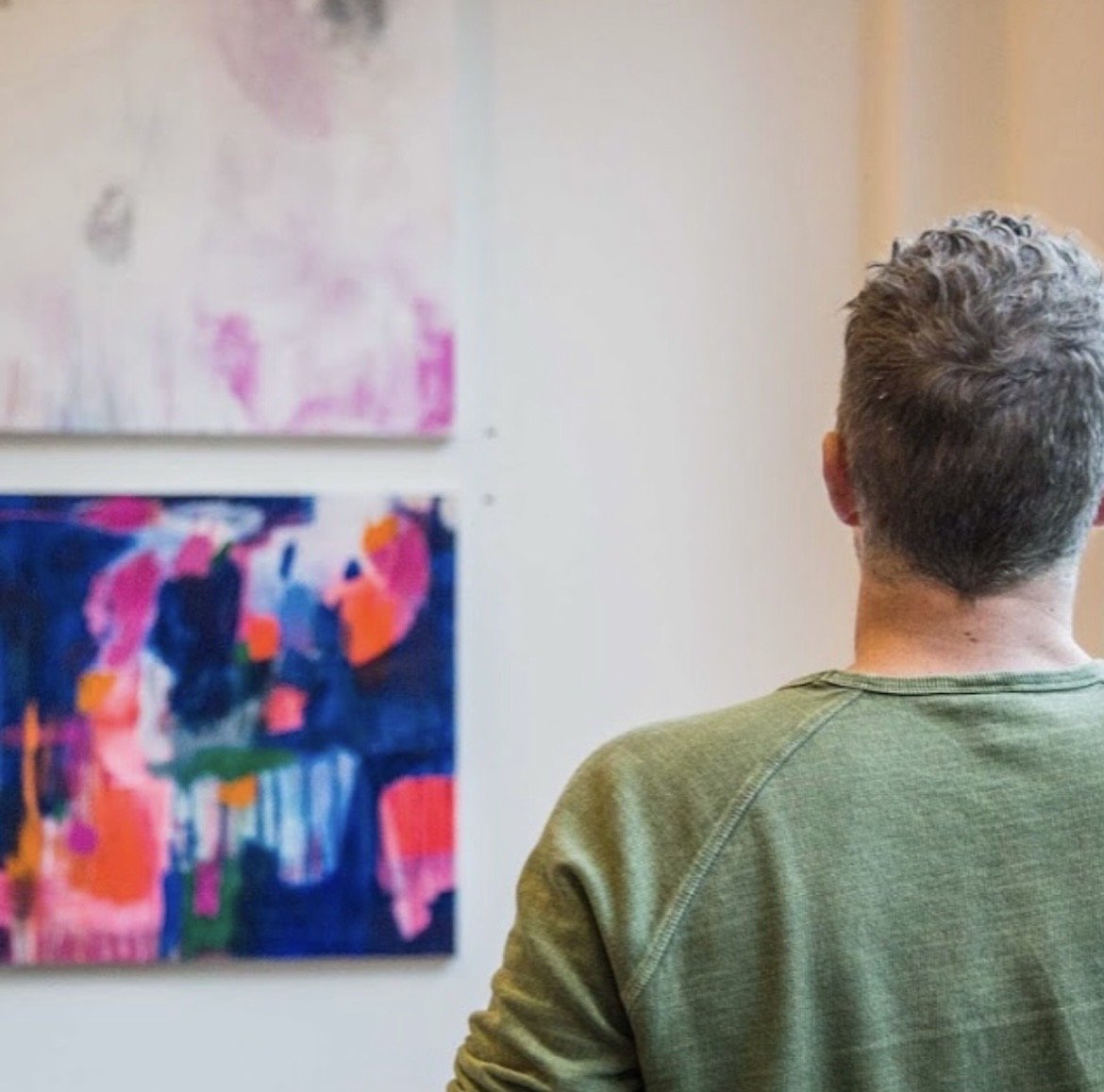 Person with gray curly hair wearing a green shirt viewing abstract art paintings on a gallery wall.