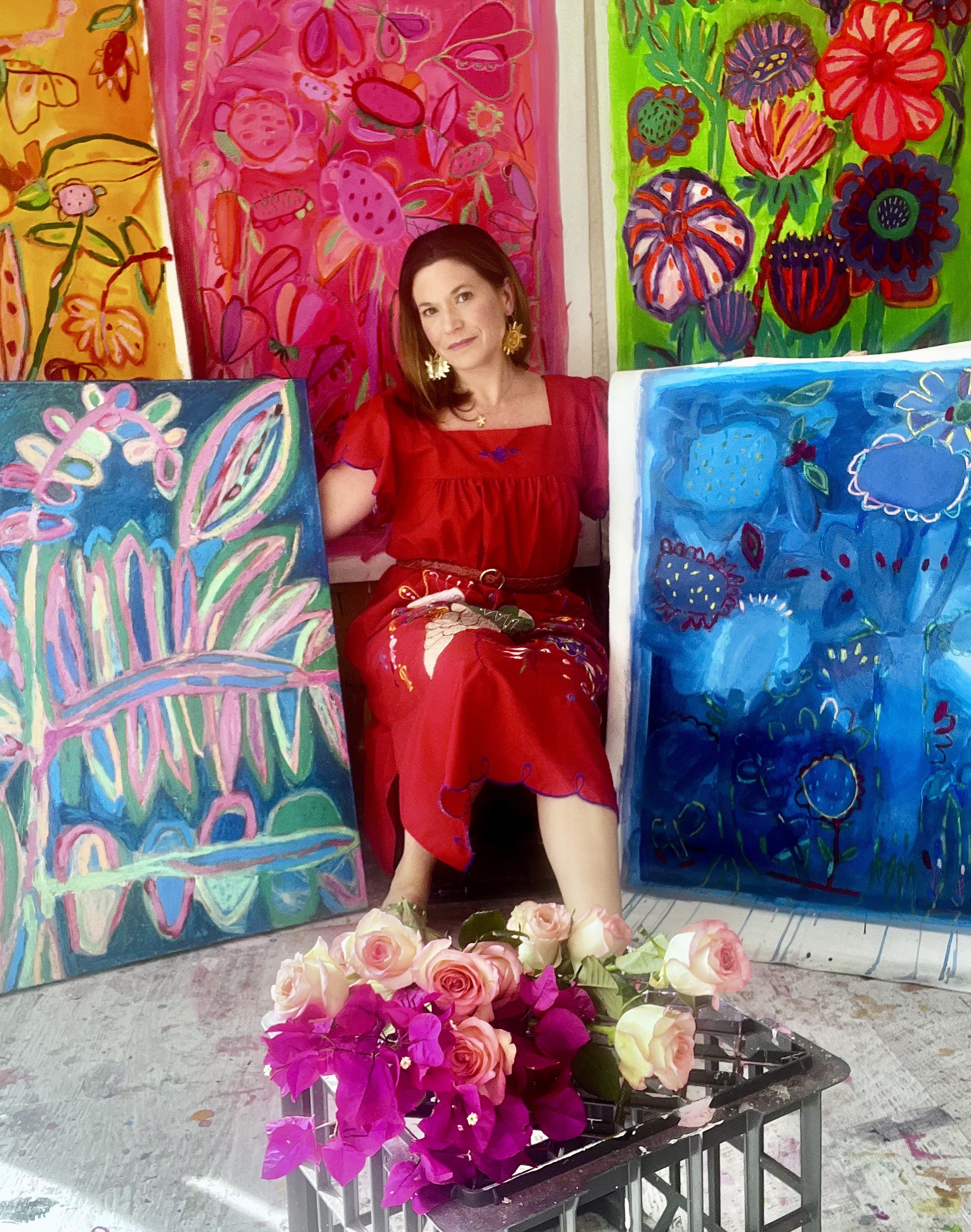 A woman with brown hair wearing a red dress with floral embroidery sitting among colorful paintings of flowers and a basket of pink and purple roses and pink bougainvillea flowers in front of her.