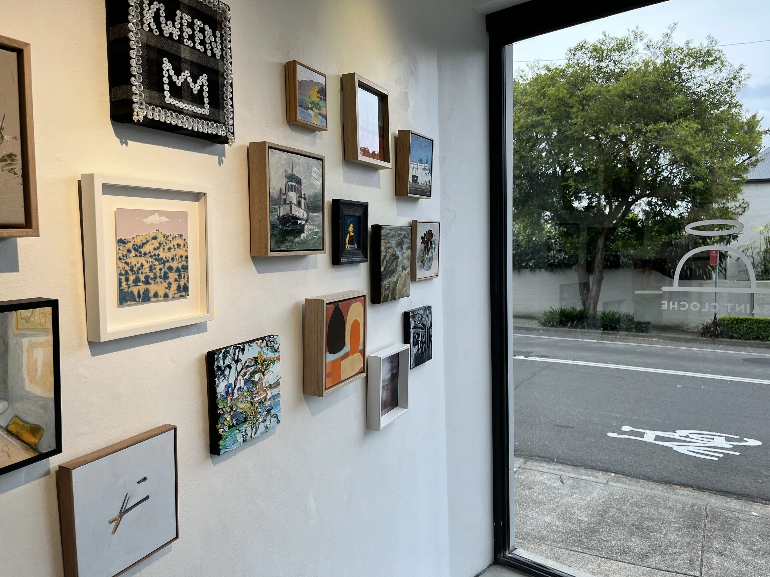 An art gallery wall displaying various framed paintings and artwork, with a large window showing an outdoor scene of trees and parked cars.