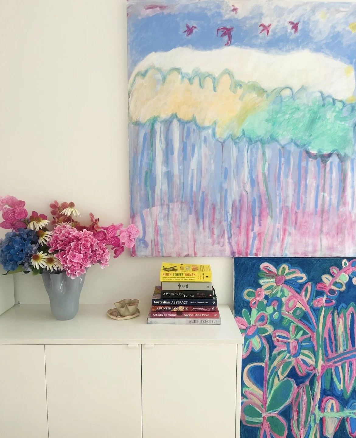 A white shelf with a vase of pink, blue, and white flowers, a small ceramic bowl, a stack of art books, and colorful abstract paintings on the wall behind.