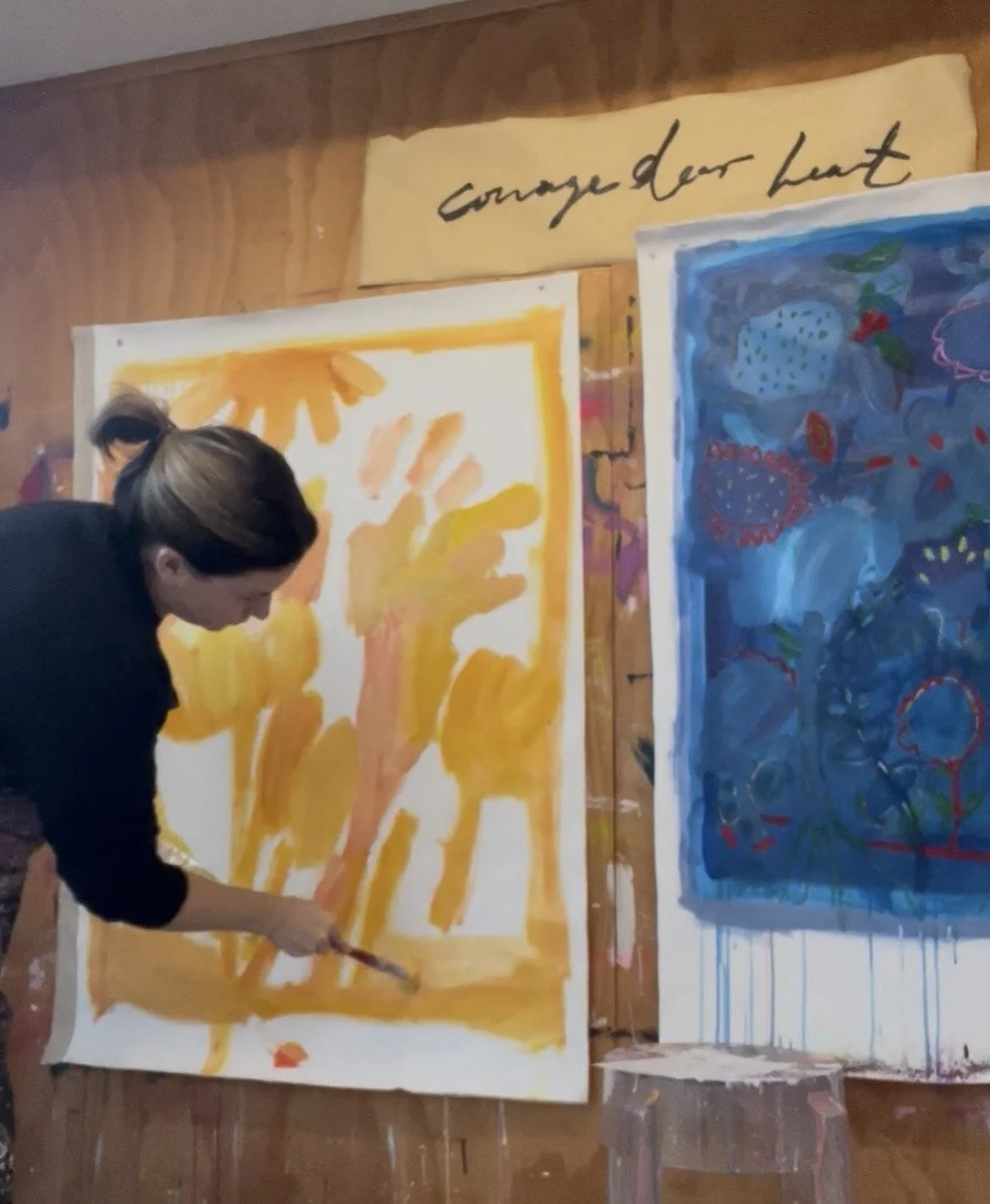 Person painting on large canvas with shades of orange and yellow, with abstract artwork featuring a hand silhouette. There are additional paintings on the wall behind. A sign above reads 'Change our Heart' in cursive.