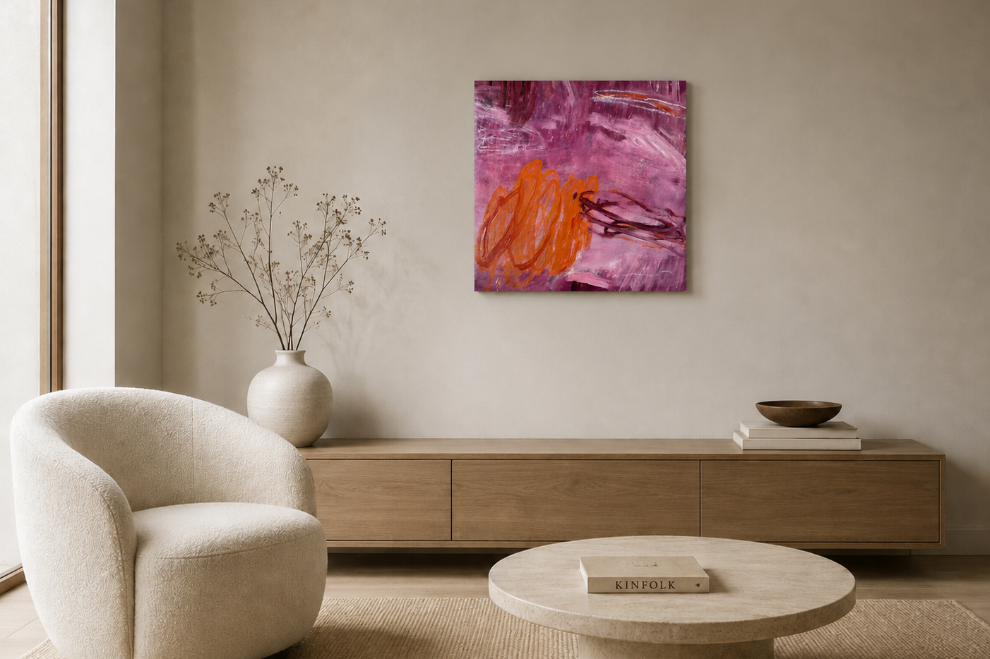 Minimalist living room with a cream armchair, a light wood sideboard, a large white vase with bare branches, a pink abstract painting on the wall, and a round cream coffee table with a book titled 'KINFOLK' on it.