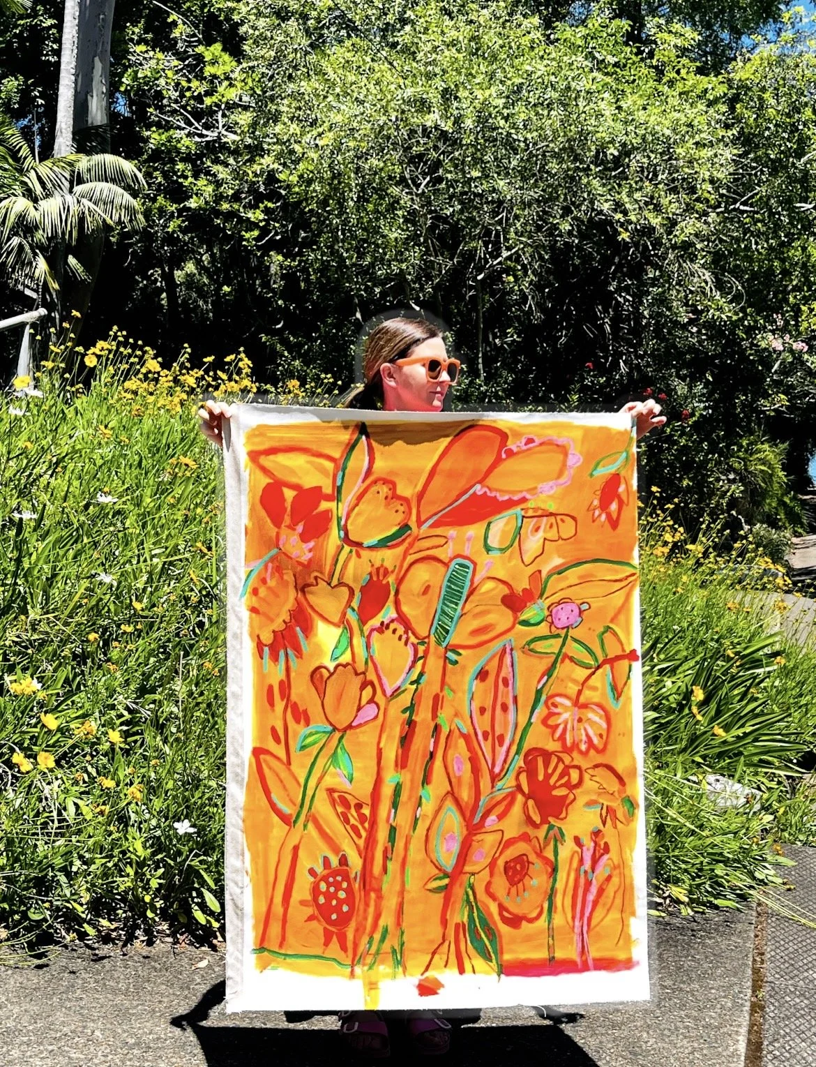 Person holding a colorful, abstract floral artwork outdoors surrounded by greenery and yellow flowers, with sunglasses and dark hair.
