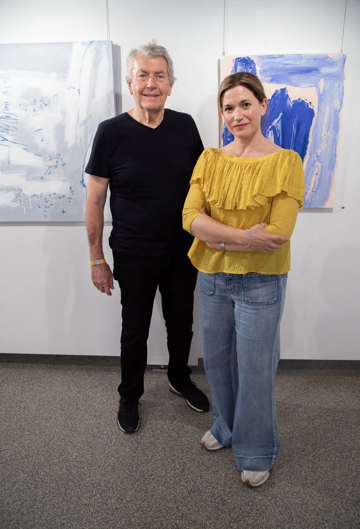 A man and a woman standing in front of abstract blue and white paintings in an art gallery. The man has gray hair and is wearing a black T-shirt and black pants. The woman has brown hair and is wearing a yellow ruffled top and blue jeans.