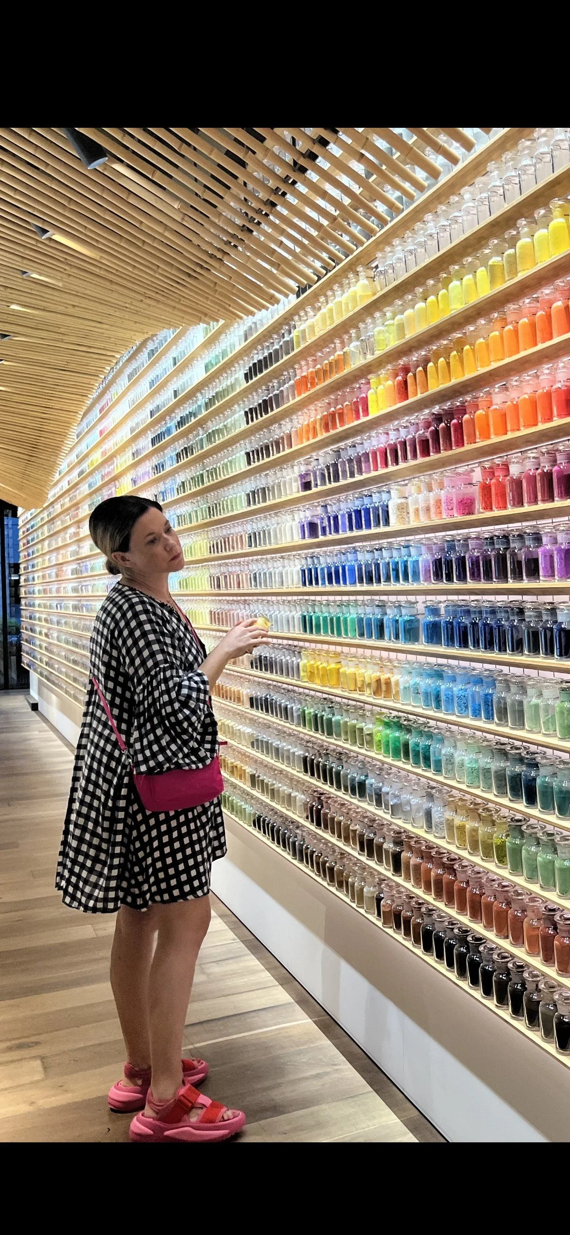 A woman in a black and white checkered dress with pink sandals and a bright pink bag is browsing a wall of colorful bottles in a modern store with wooden ceiling design.