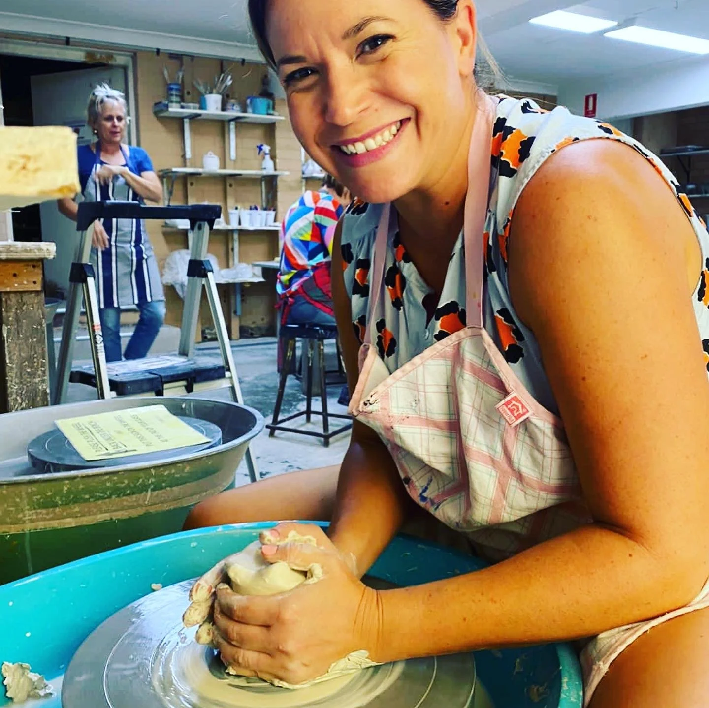 Woman smiling and kneading dough on a circular table, in a pottery studio with shelves and another person in the background.