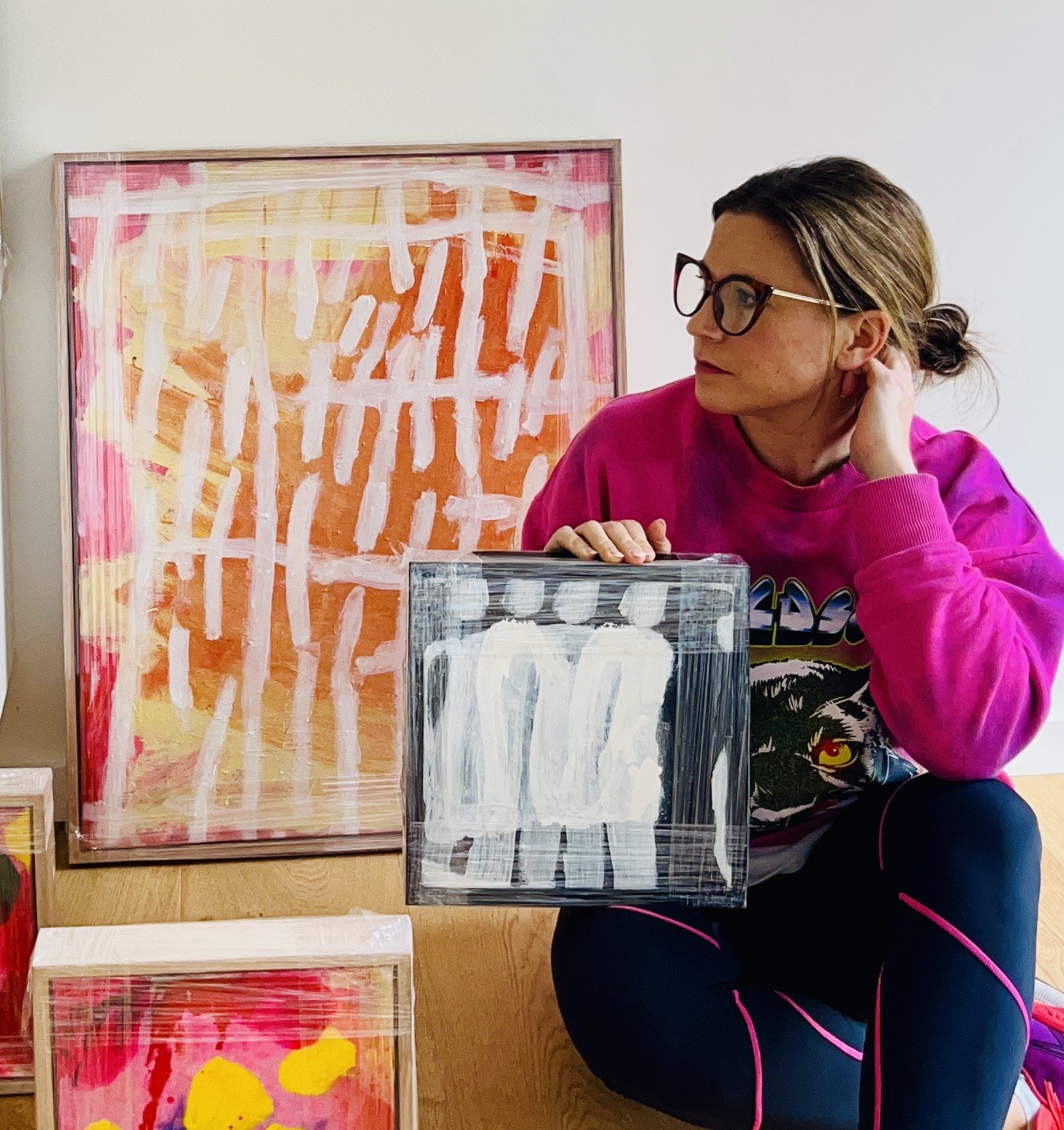 A woman with glasses wearing a pink sweatshirt sitting beside three abstract paintings on display.