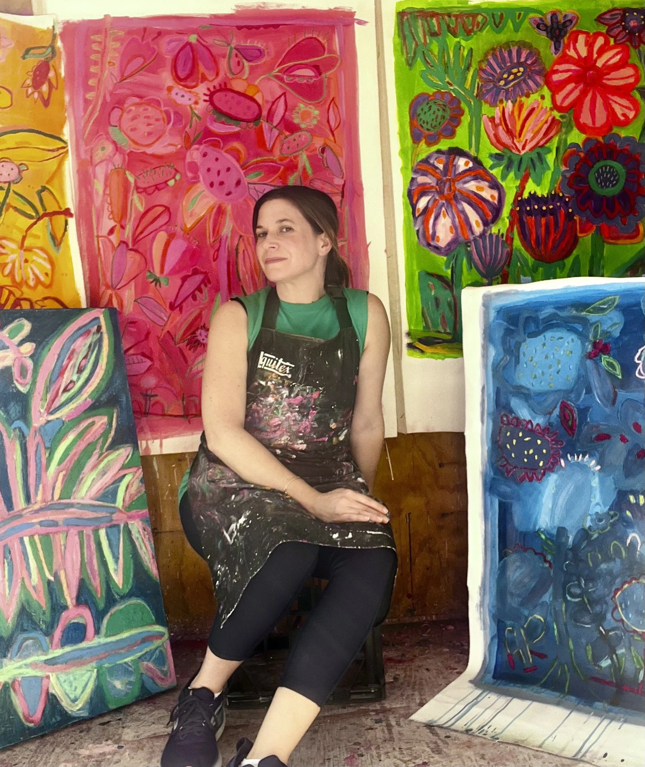 A woman with brown hair, wearing a green shirt and a black apron, sitting among colorful paintings of flowers and butterflies in an art studio.