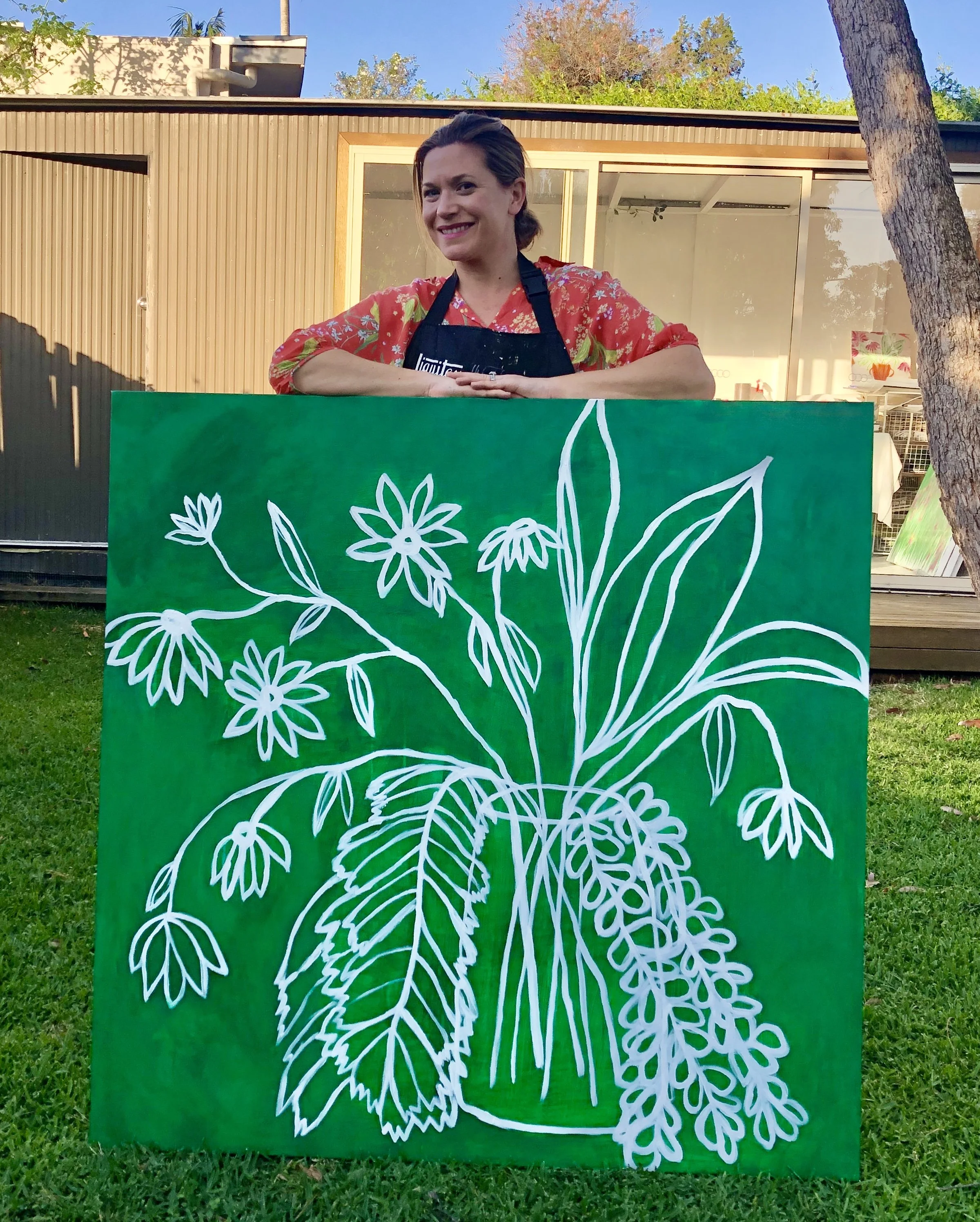 Woman smiling behind a green sign with white floral design, outdoors in front of a wooden building, trees, and a blue sky.