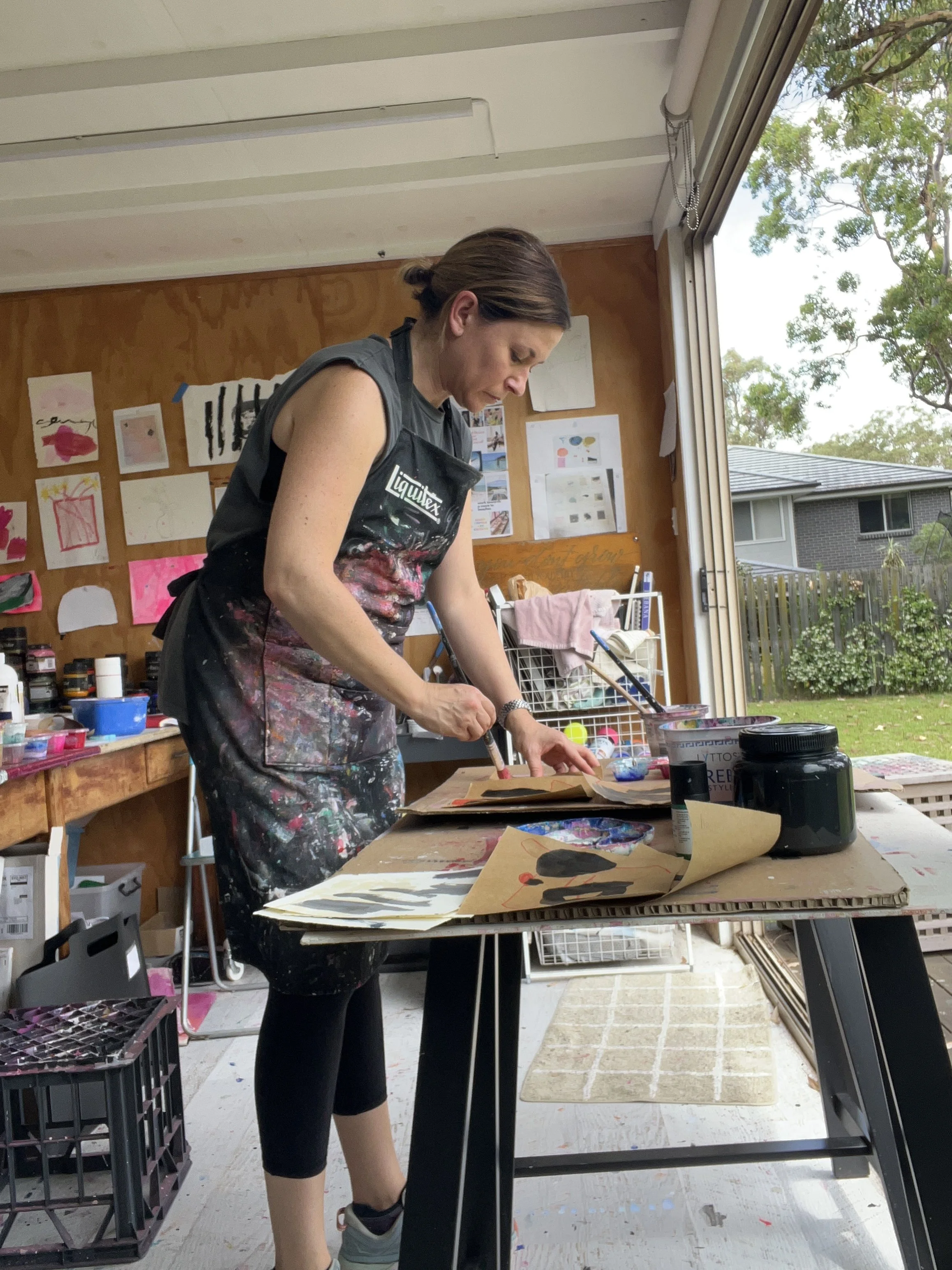 A woman with brown hair tied back, wearing a black apron splattered with paint, is working on art projects at a table in a brightly lit studio, with artwork and supplies around her and a large open window showing trees and a house outside.