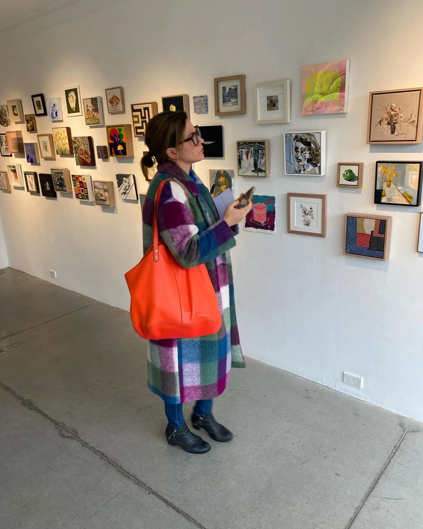 Woman in colorful plaid coat with orange bag looking at artwork on gallery wall.