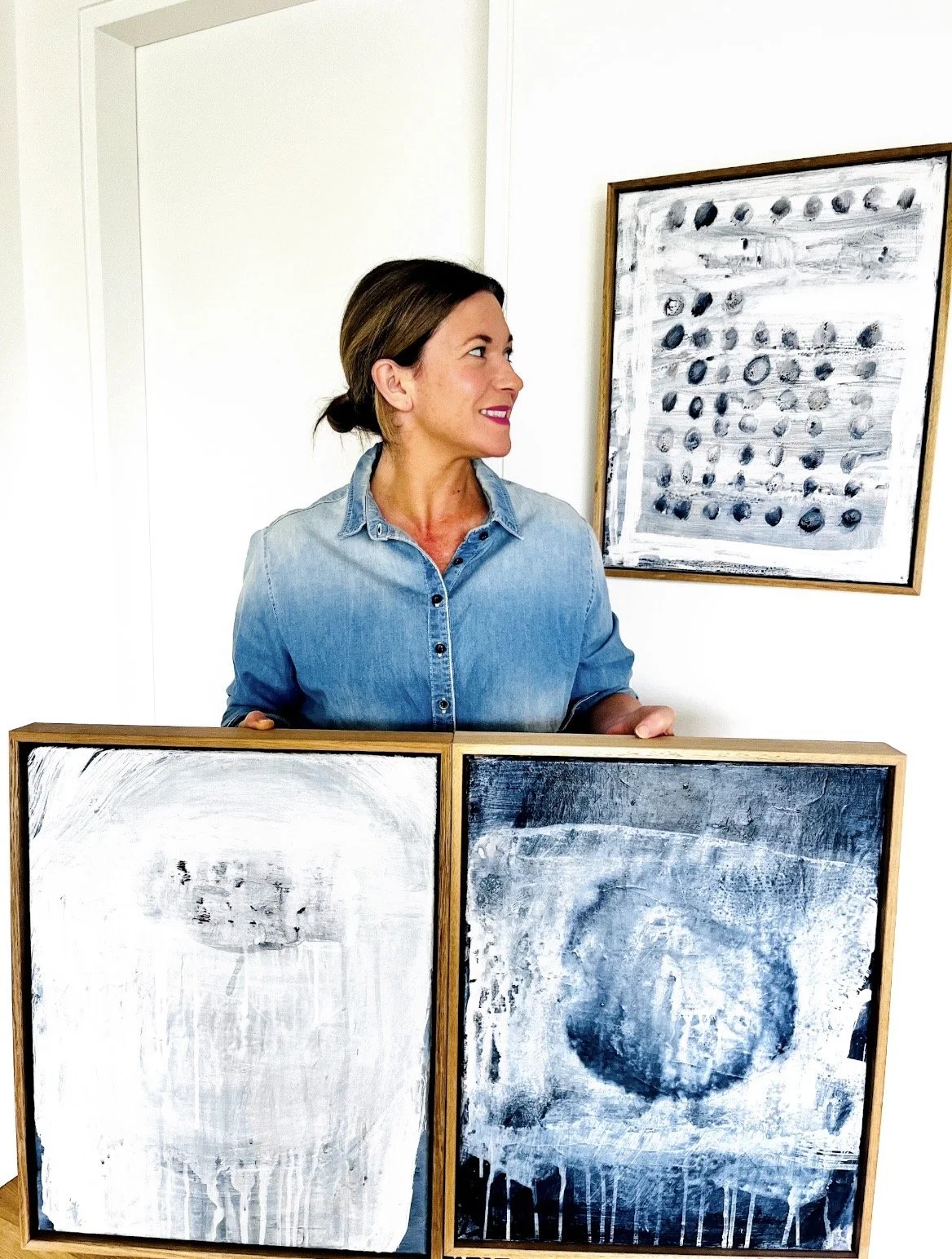 Woman in denim shirt holding two abstract black-and-white paintings in frames, standing in front of a third framed artwork on a white wall.