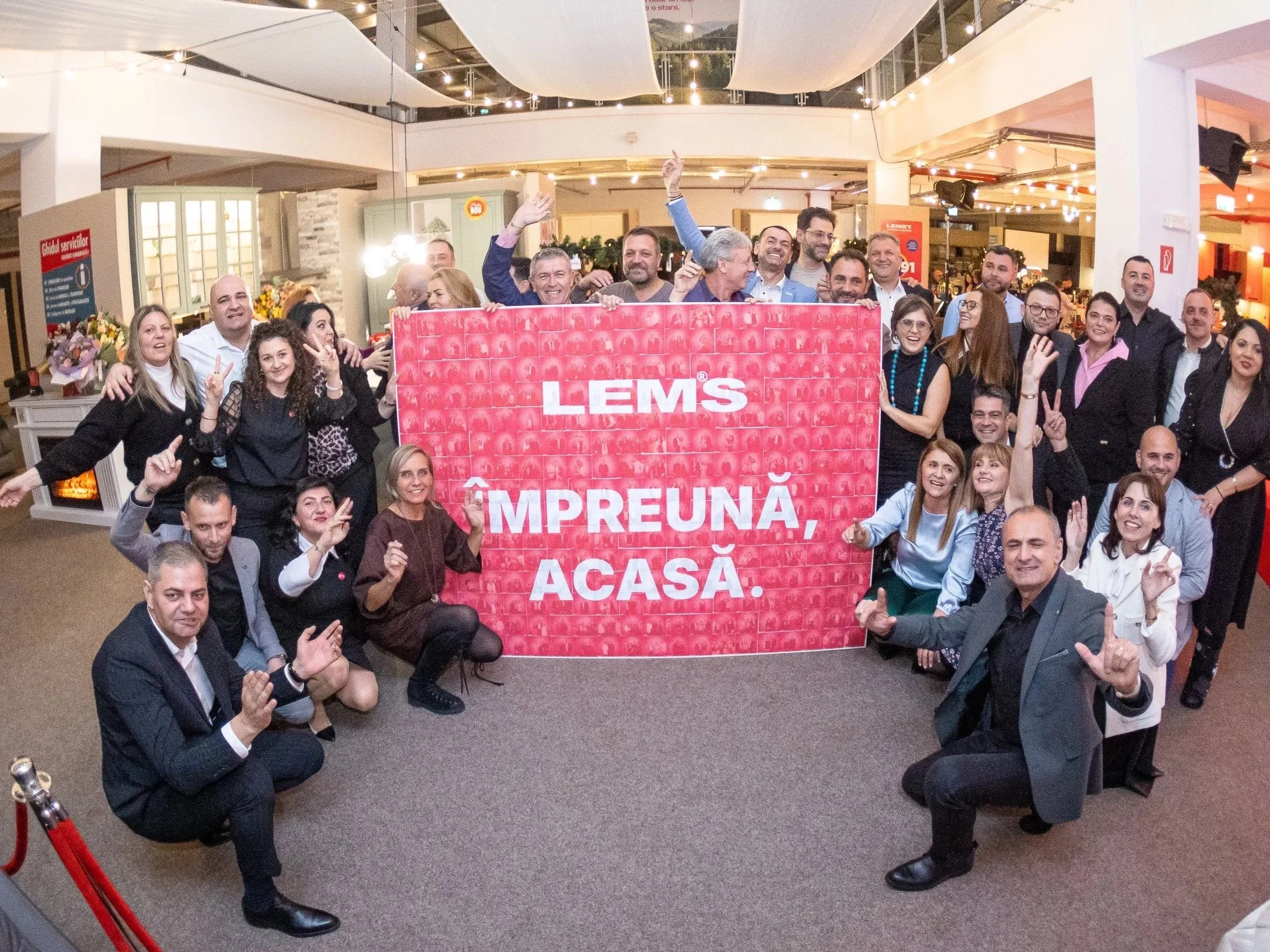 A group of people gathered indoors, smiling and posing for the photo. They are holding a red banner with white text that reads "LEMS IMPREUNĂ, ACASĂ." There are string lights hanging from the ceiling and a few decorative elements in the background.