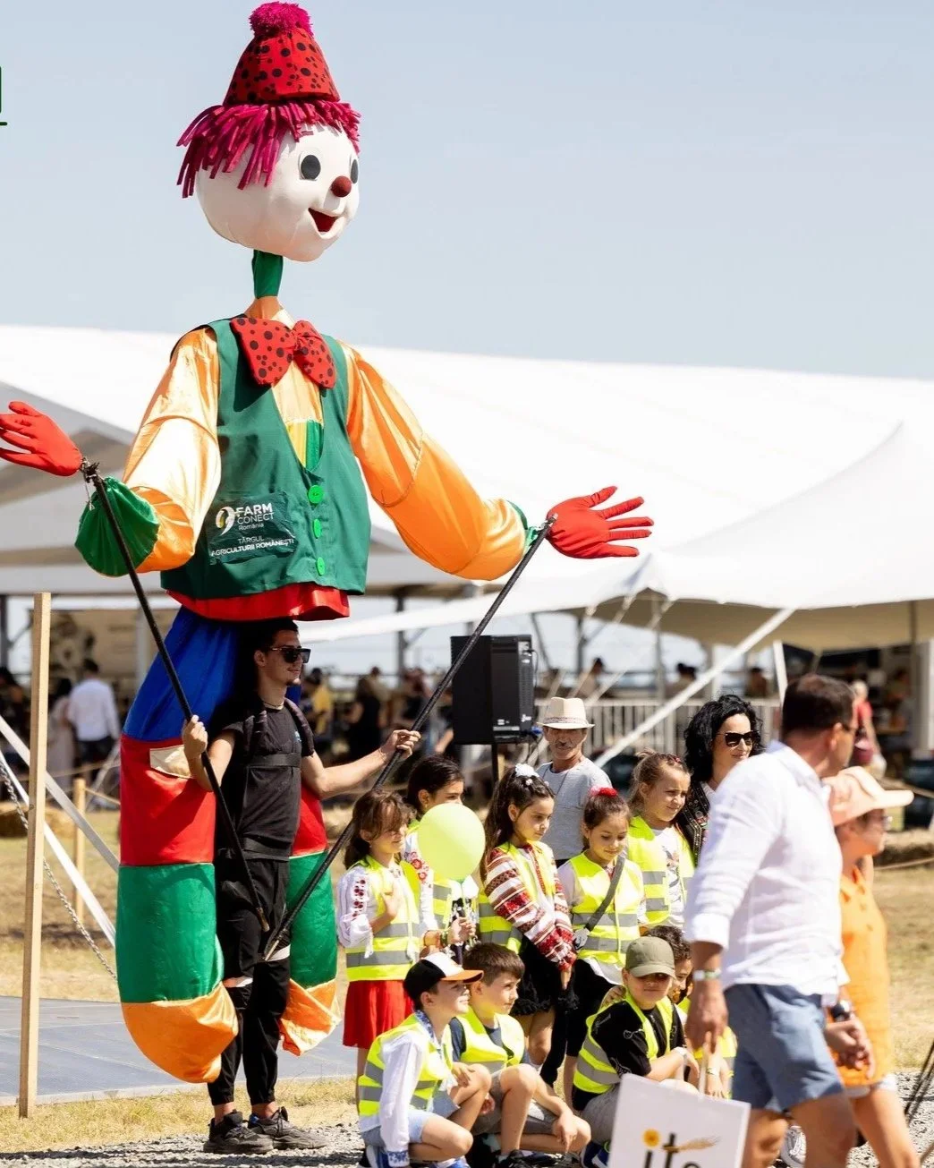 A giant clown puppet with a smiling face, red hair, and a party hat, standing above a group of children and adults at an outdoor event.