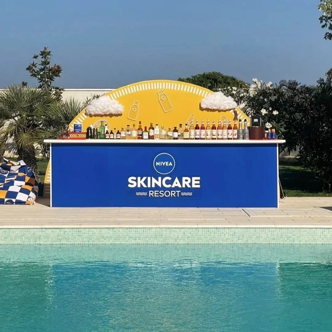 A poolside bar at a skincare resort, with a large blue sign reading 'Nivea Skincare Resort' and a yellow backdrop decorated with clouds and bottles.