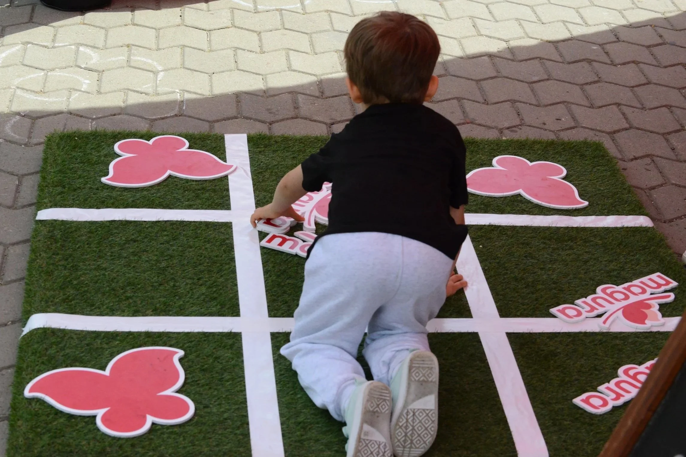 Two children, a girl and a boy, are playing with pink floral cutouts on a grassy surface enclosed by tape, with a paved walkway in the background.