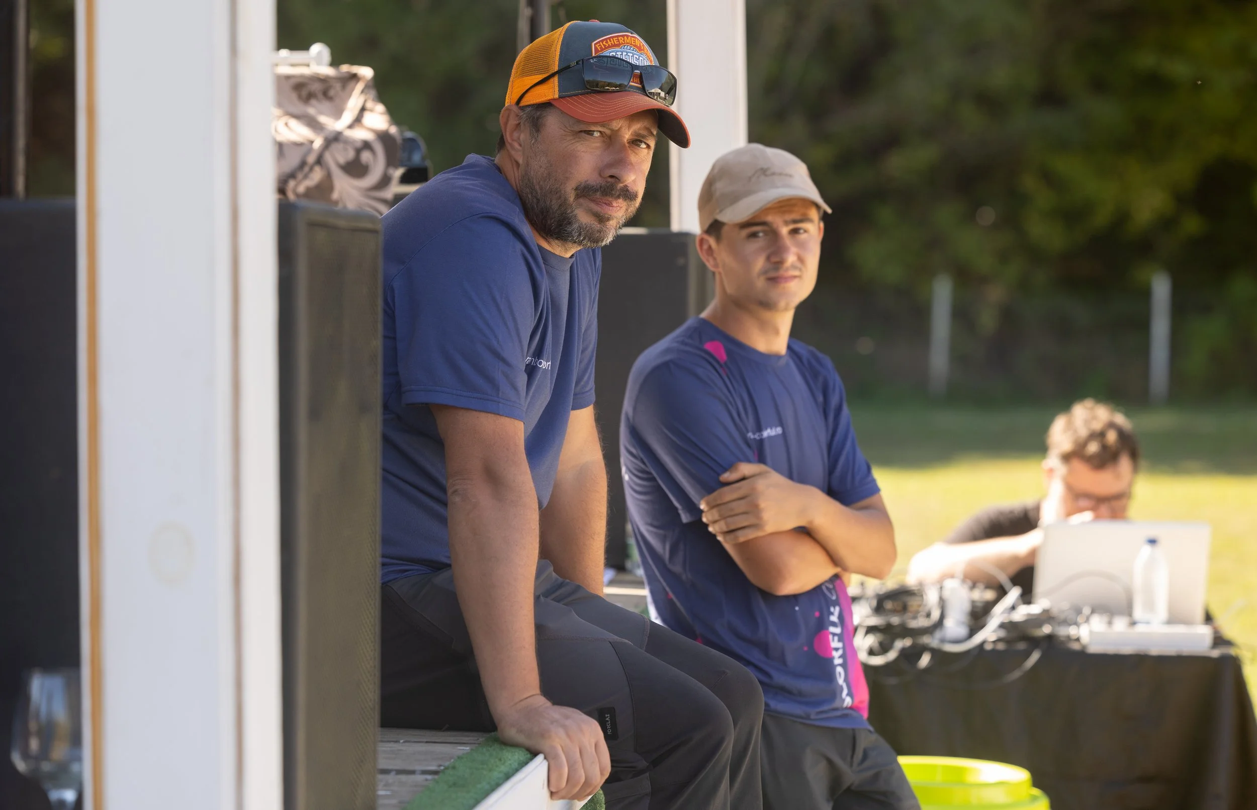 Two men sitting outdoors, with trees in the background. One man is wearing a blue shirt, an orange and blue cap, and sunglasses on his head, looking towards the camera. The other man is dressed in a blue shirt and a beige cap, crossing his arms and l