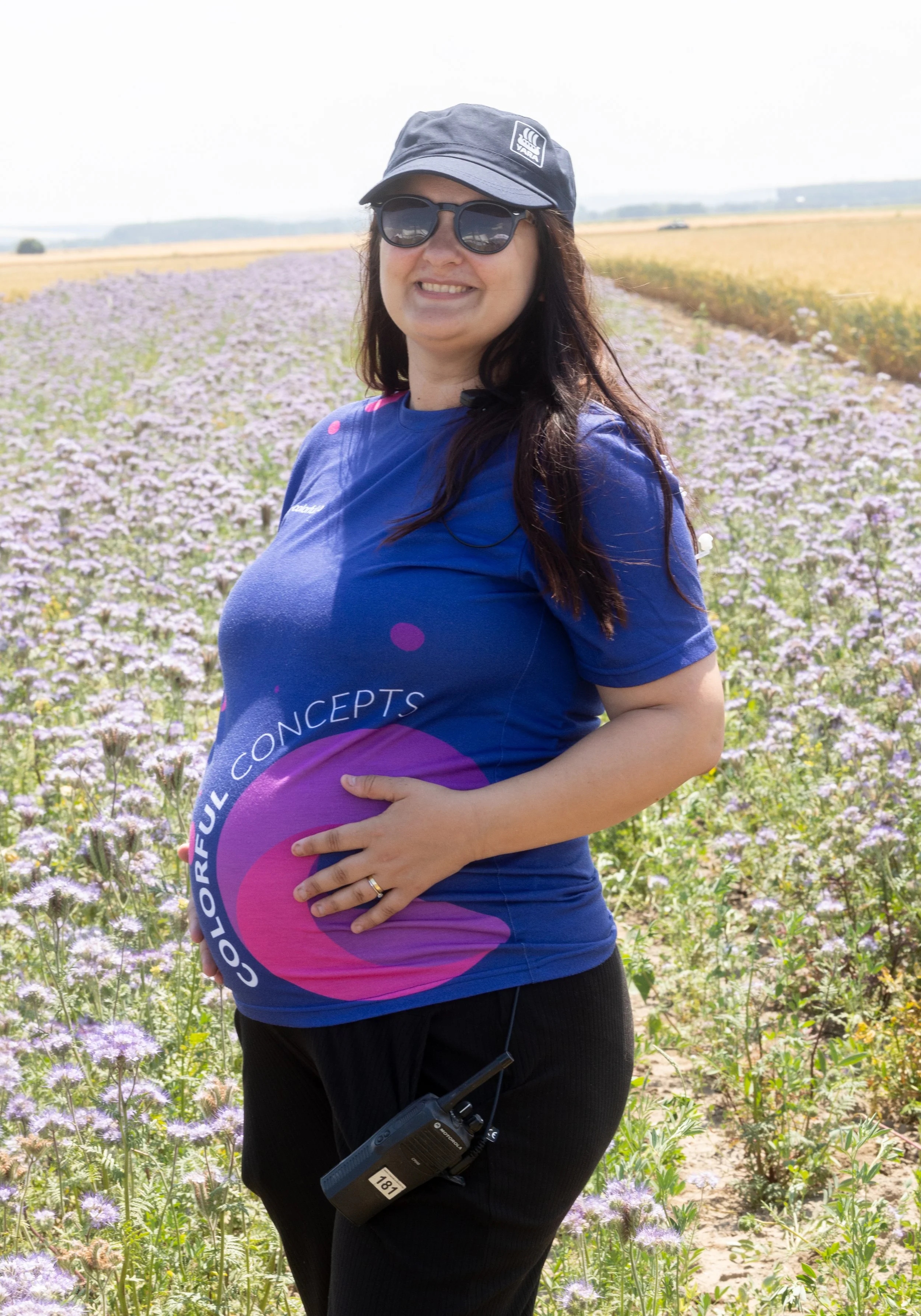 Woman in sunglasses and a cap standing in a field of purple flowers, holding her pregnant belly, with a walkie-talkie hanging from her waist.