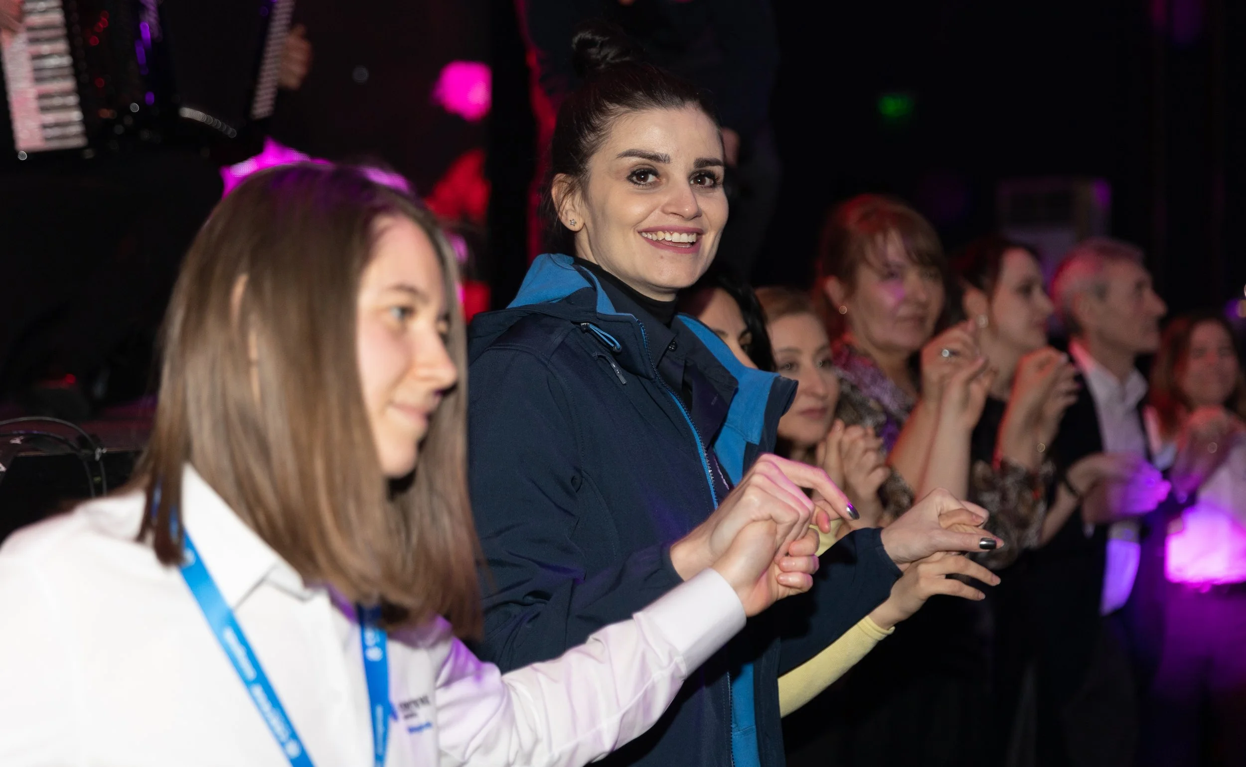 Group of people sitting in an audience, smiling and clapping at an event with colorful lighting.