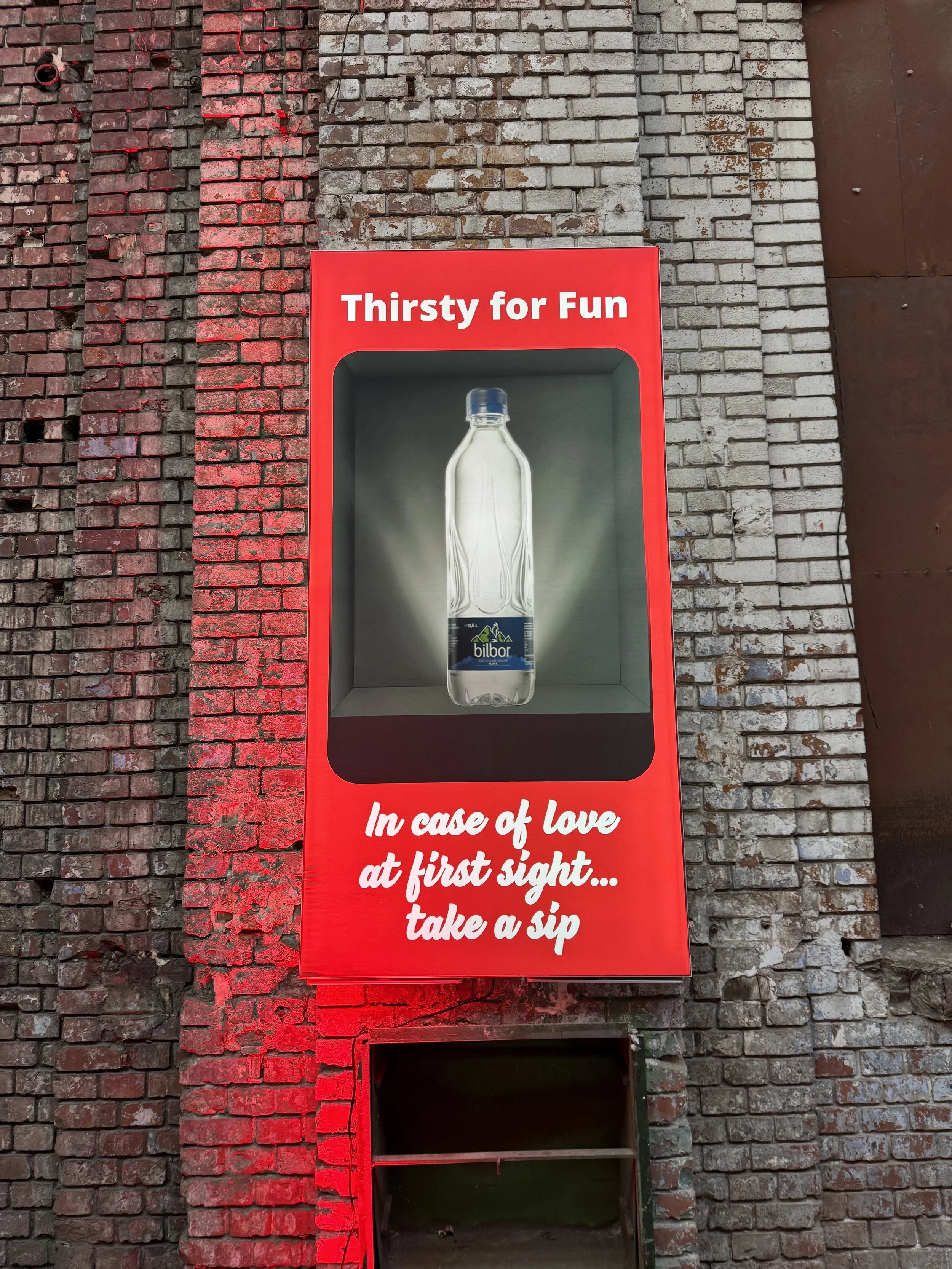 A red vending machine for bottled water with the words "Thirsty for Fun" at the top and the phrase "In case of love at first sight... take a sip" at the bottom, mounted on a weathered brick wall.