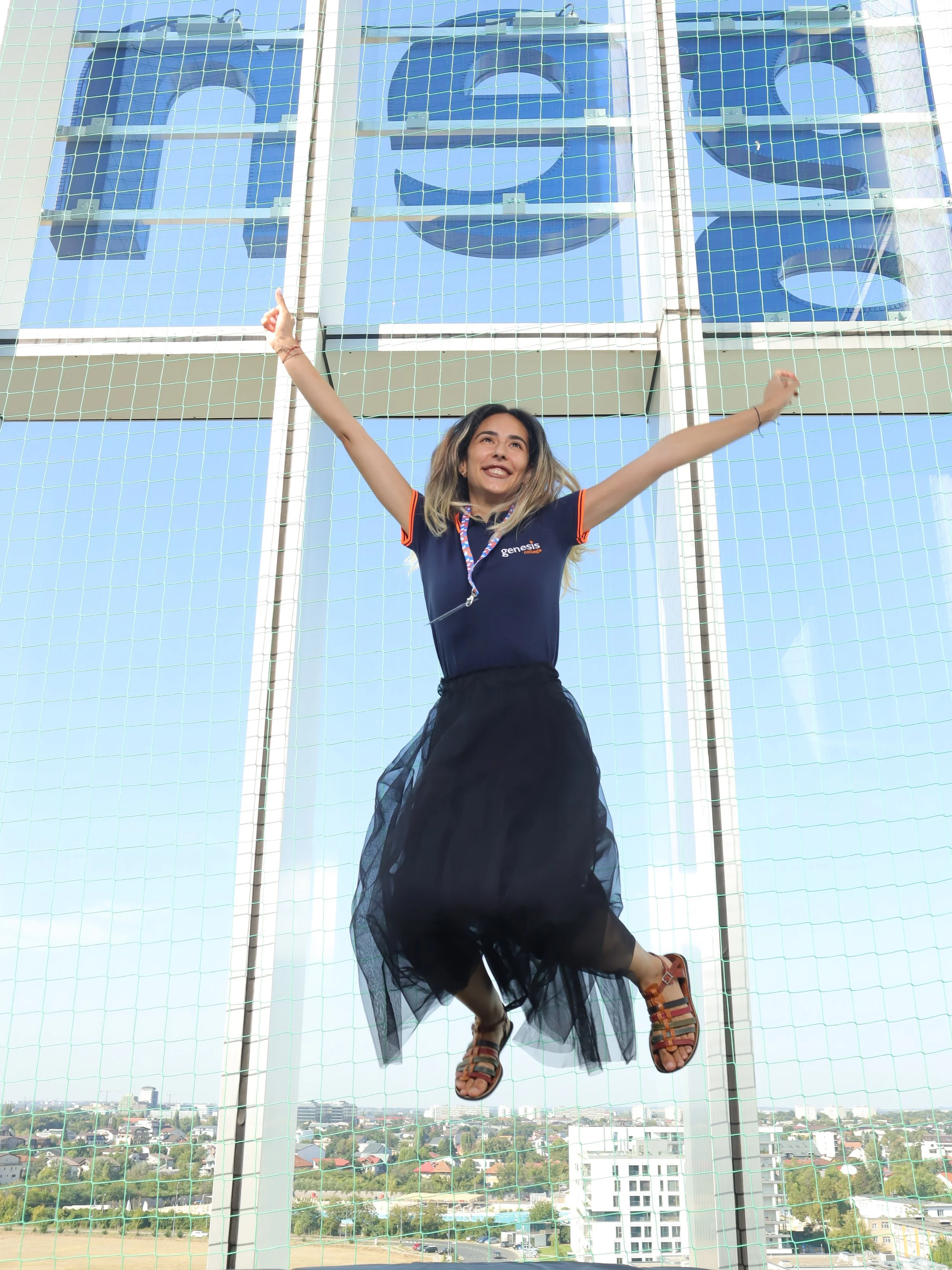 A woman in a black skirt, colorful sandals, and a gray Genesis College sports uniform is standing behind a sports net, with her arms raised and smiling. Behind her, a large building with the Genesis logo and a blue sky are visible.