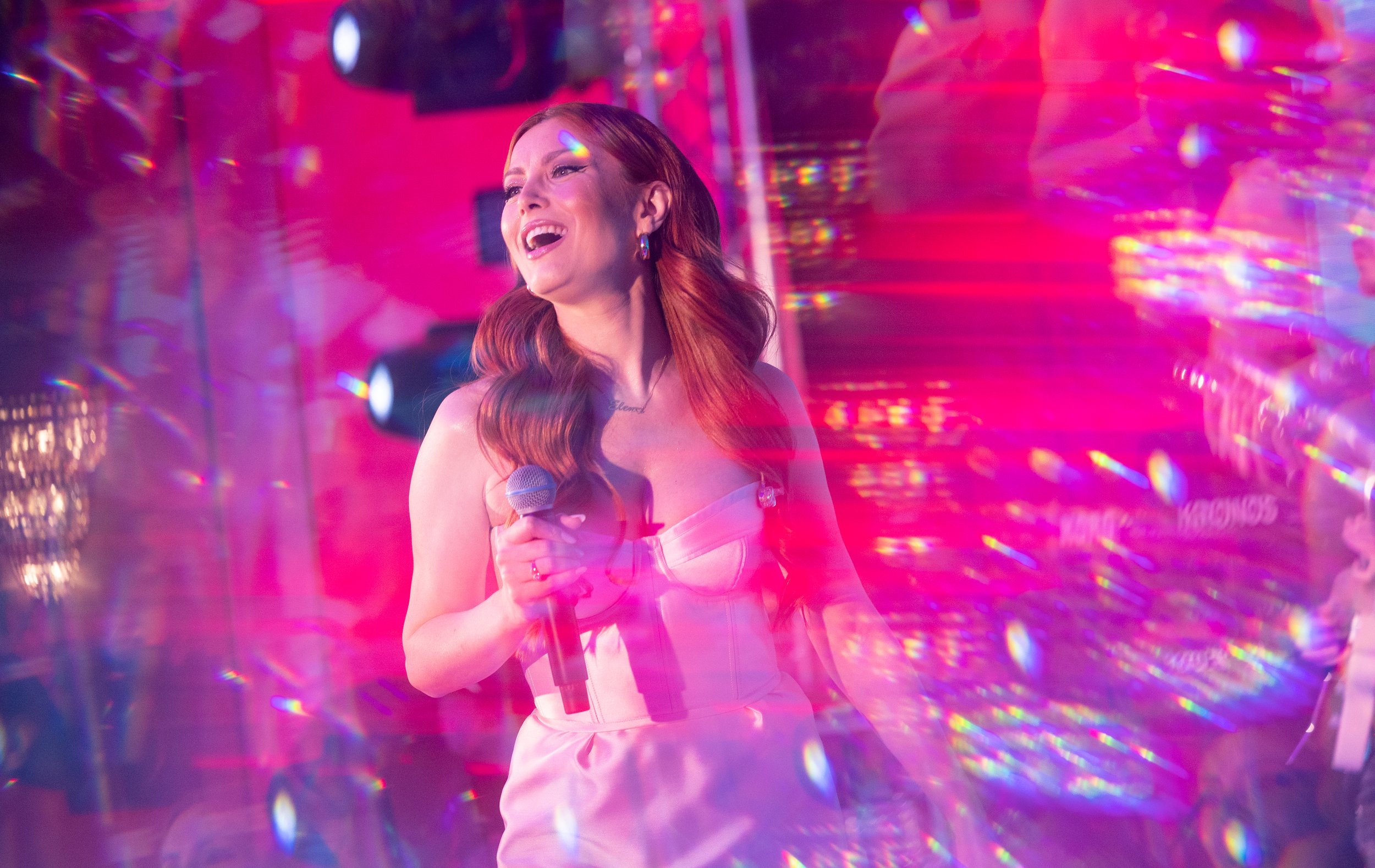 A woman with long red hair wearing a pink dress, holding a microphone and singing in a colorful, neon-lit club.