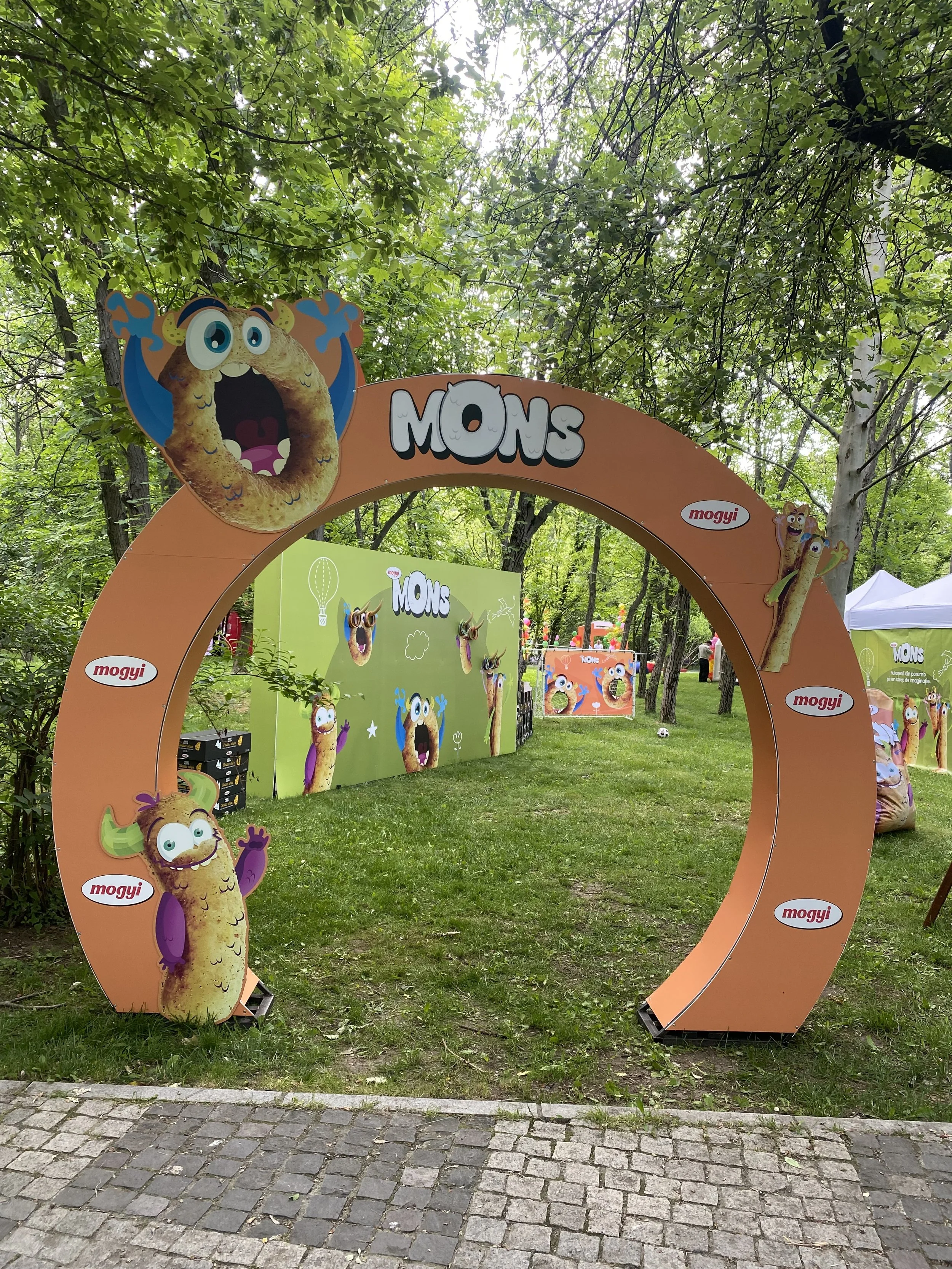 Colorful outdoor display with cartoon monster characters and the word 'MOMS' on a large orange circular frame, set in a green, wooded area.