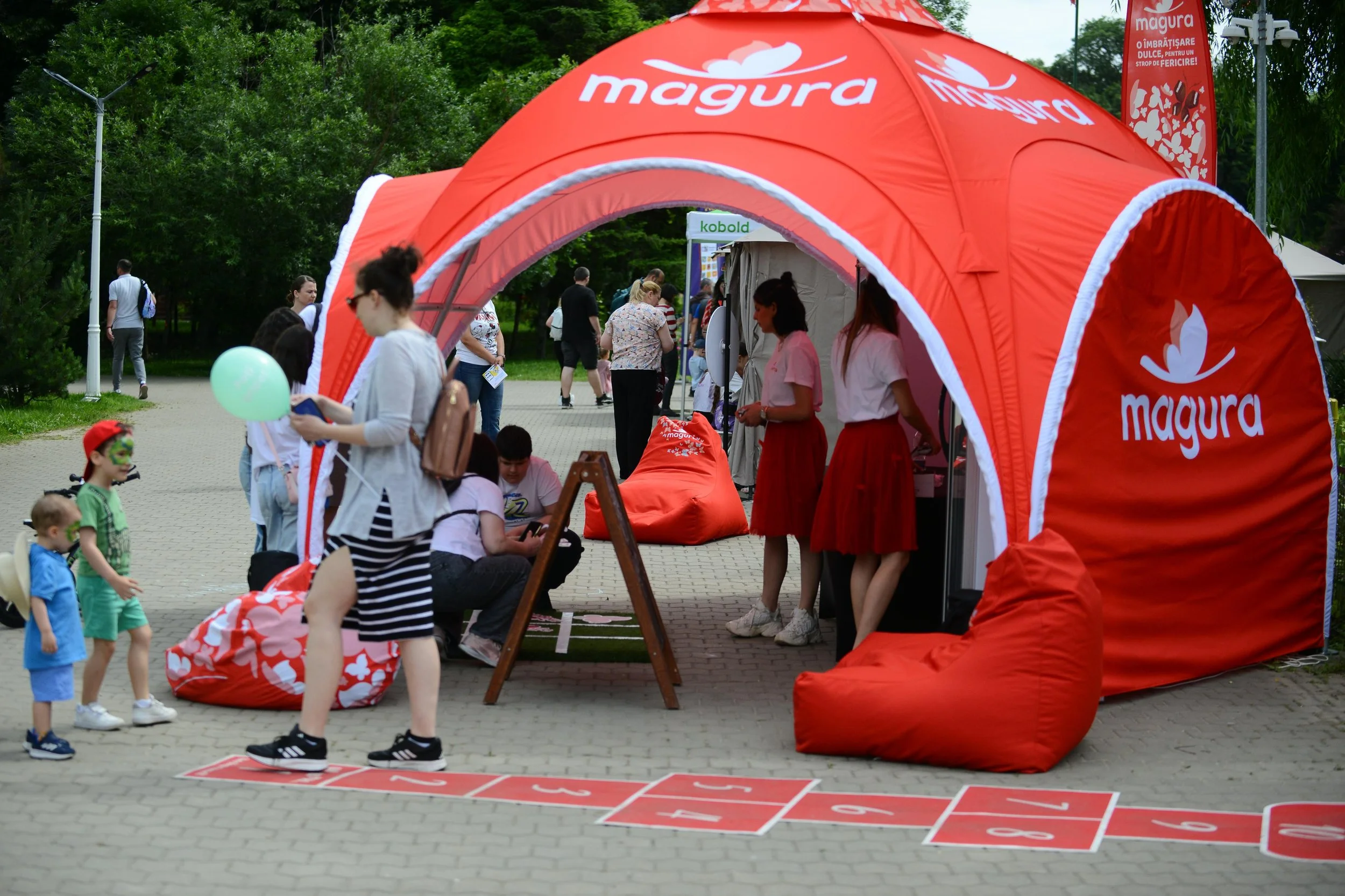 A crowded outdoor event with people walking and standing under a large red tent with the word 'Magura' printed on it. There are children and adults, some engaging in activities like looking at their phones, and others waiting in line or talking. A gr